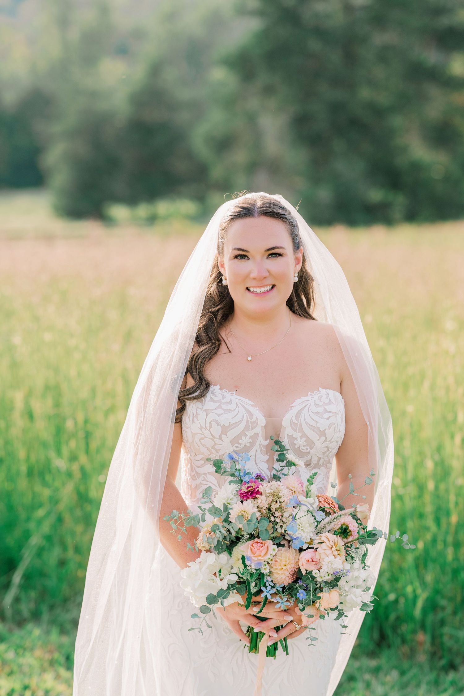 Smiling bride in strapless lace gown and veil holds colorful bouquet in sunlit green field.