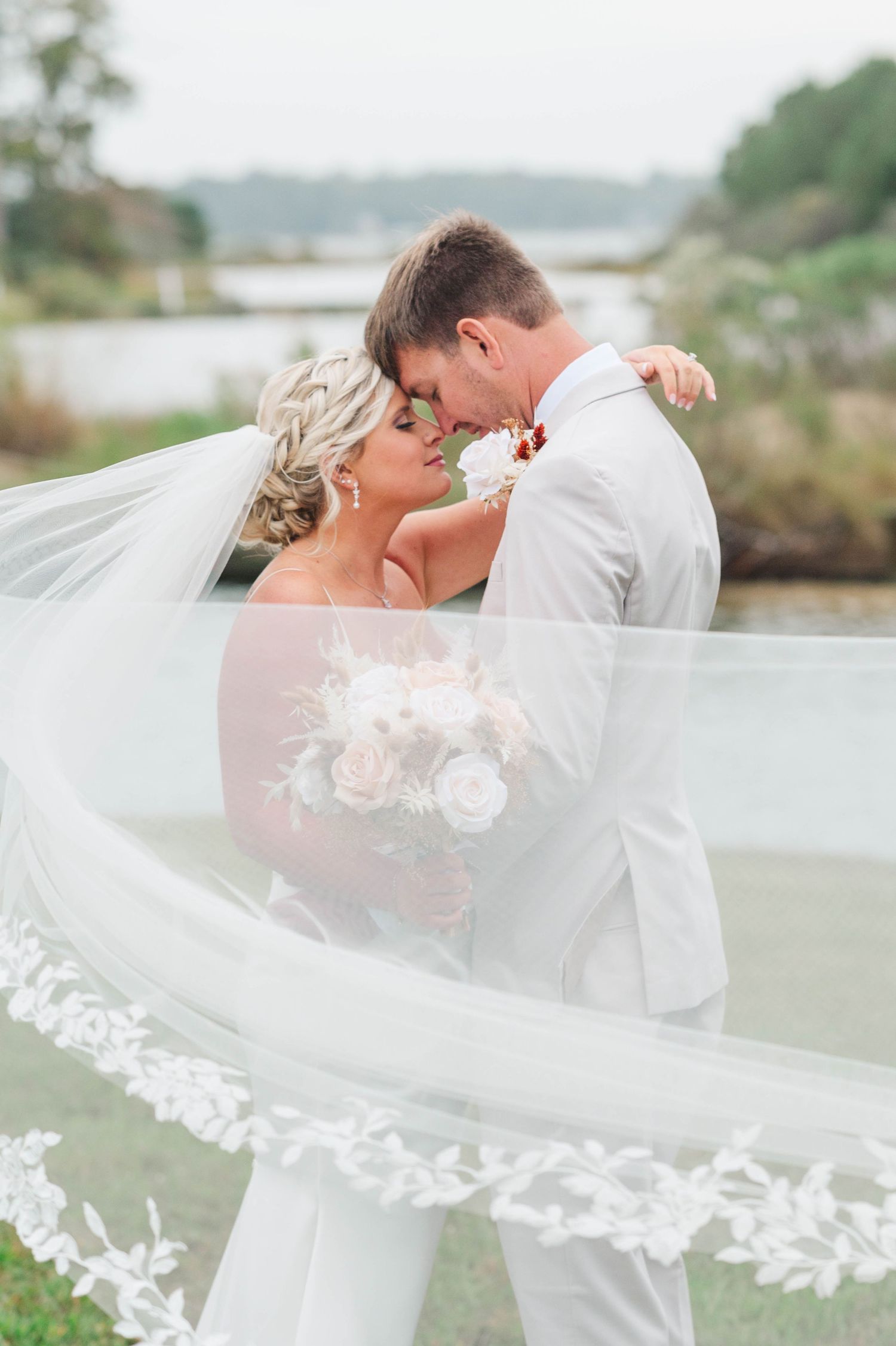 Romantic wedding couple embraces under flowing white veil with delicate floral lace details in outdoor setting.