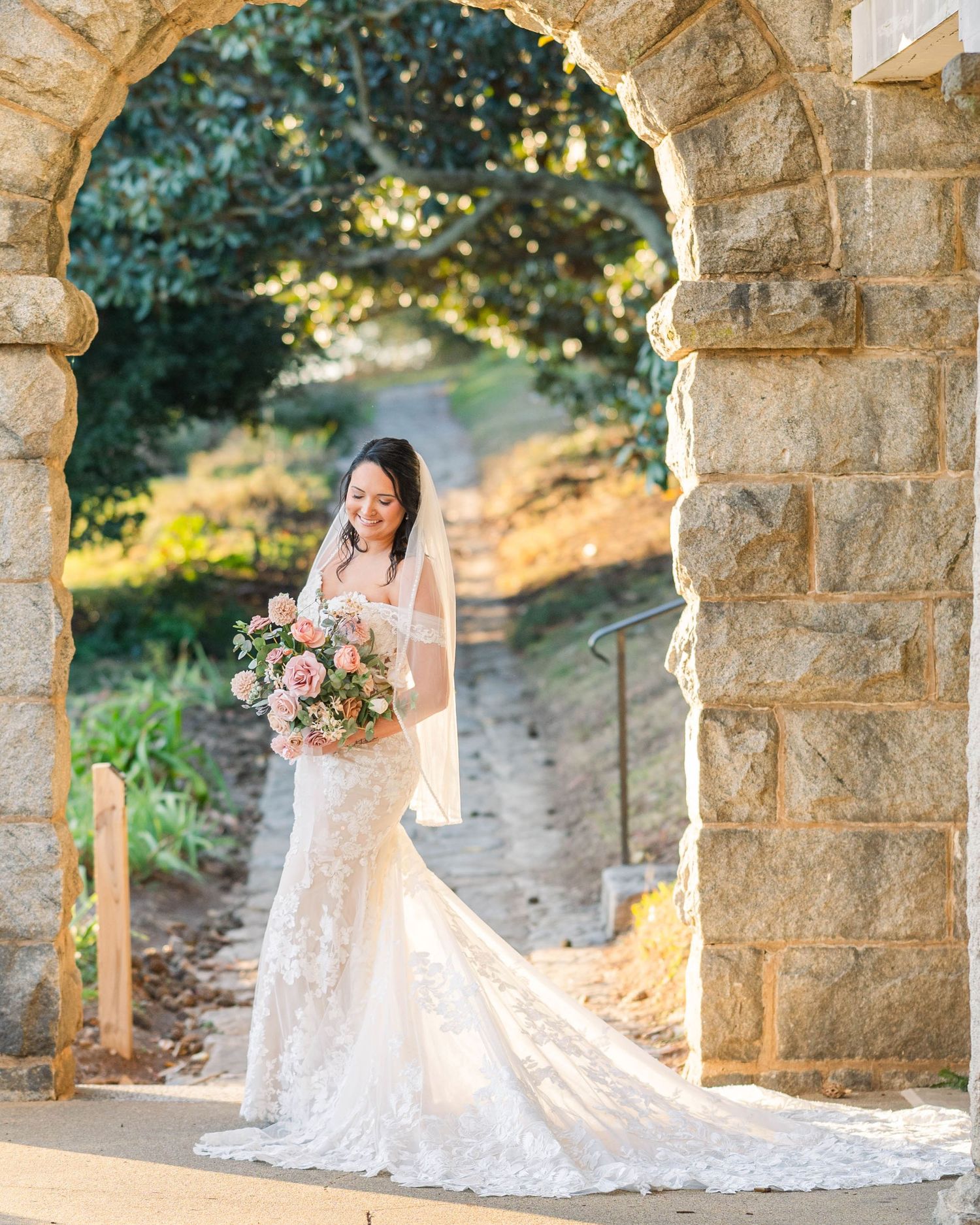 A bride in a flowing white lace gown poses under a stone archway during golden hour at Maymont Park, VA. Krista Alyse Photography