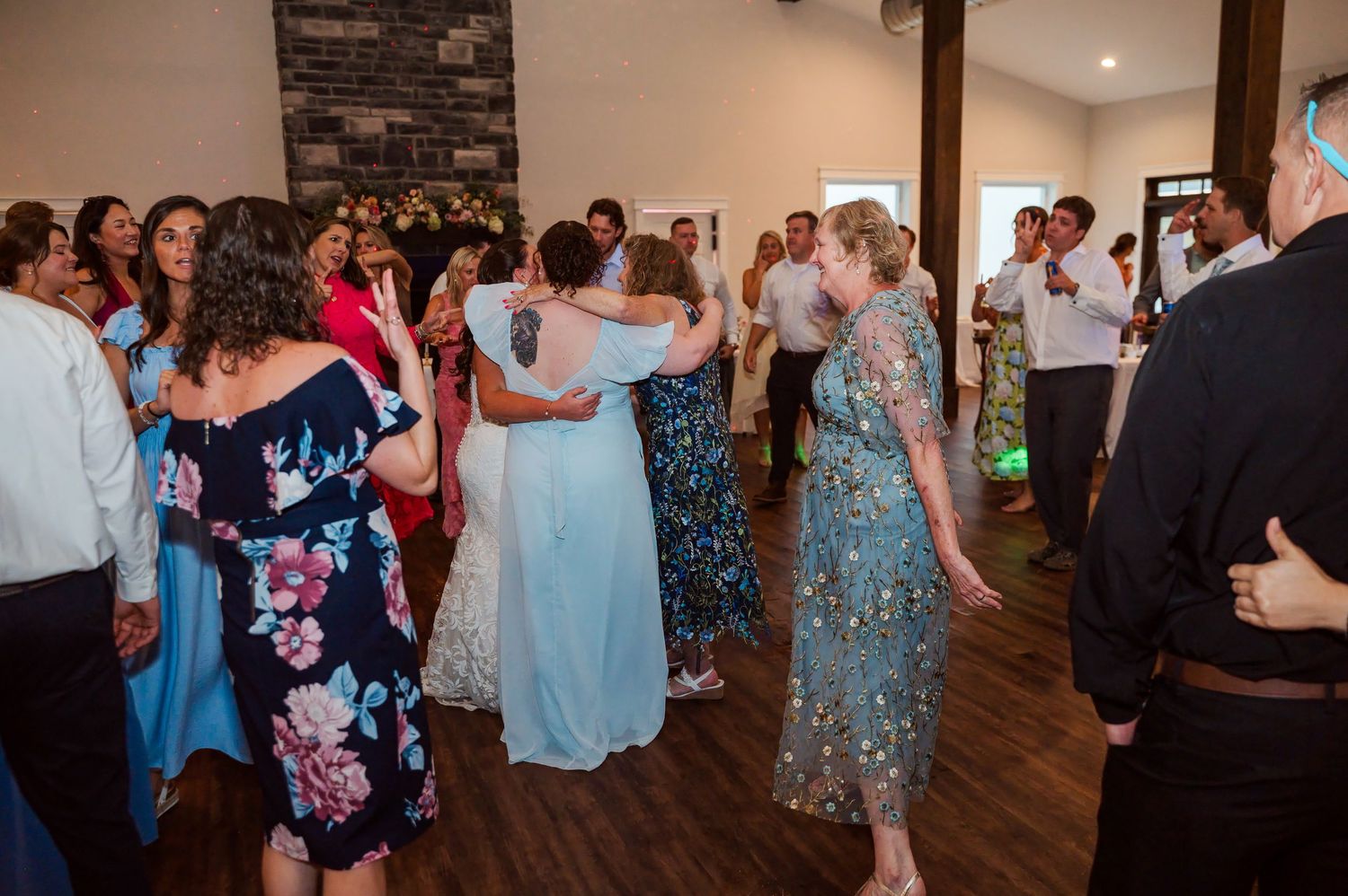 Wedding guests in formal dresses and suits dance and celebrate together on a wooden dance floor inside a rustic reception venue.