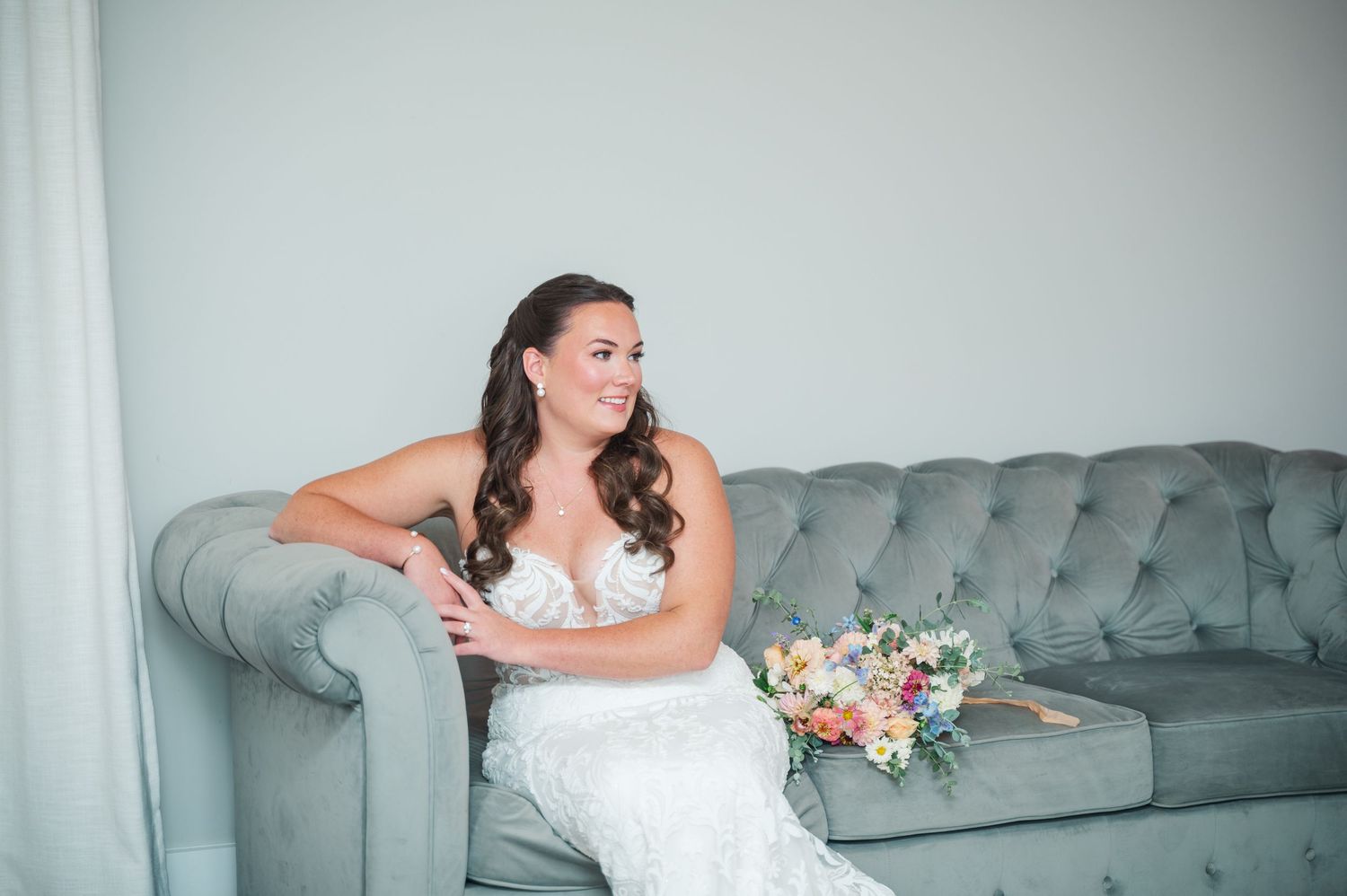Bride in lace wedding gown sits on gray tufted sofa holding colorful bouquet, smiling elegantly.