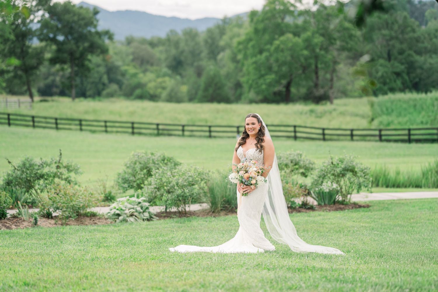 Bride in white gown and veil holds bouquet on lush green lawn with fence and mountains backdrop.