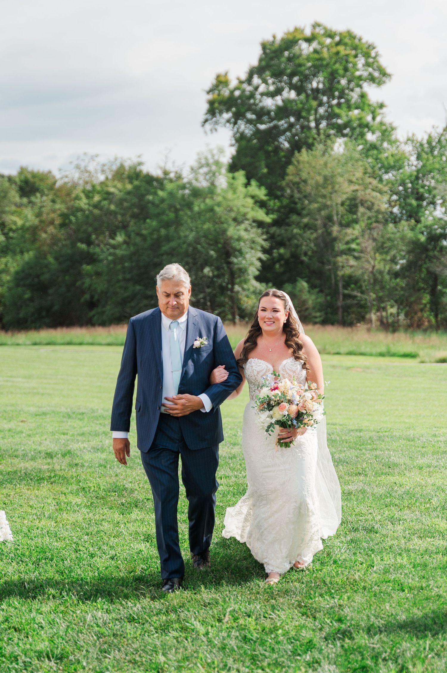 Smiling bride in lace gown walks down the aisle with her father during an outdoor wedding ceremony.