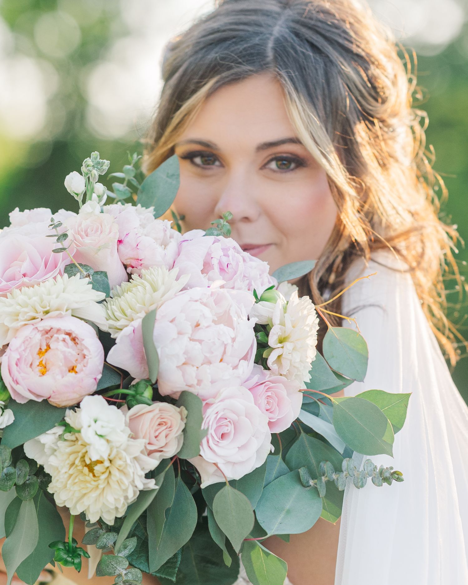 Bride holding a lush bouquet of pink peonies and roses with greenery, wearing a white dress and soft bridal makeup.