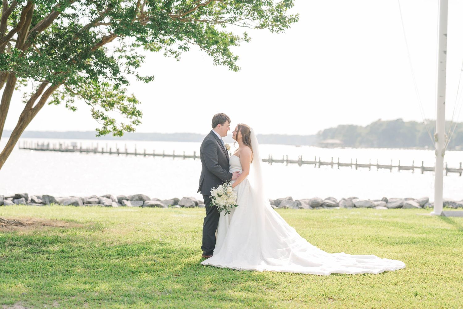 Romantic wedding couple shares a tender moment under a tree by the waterfront on a sunny day.