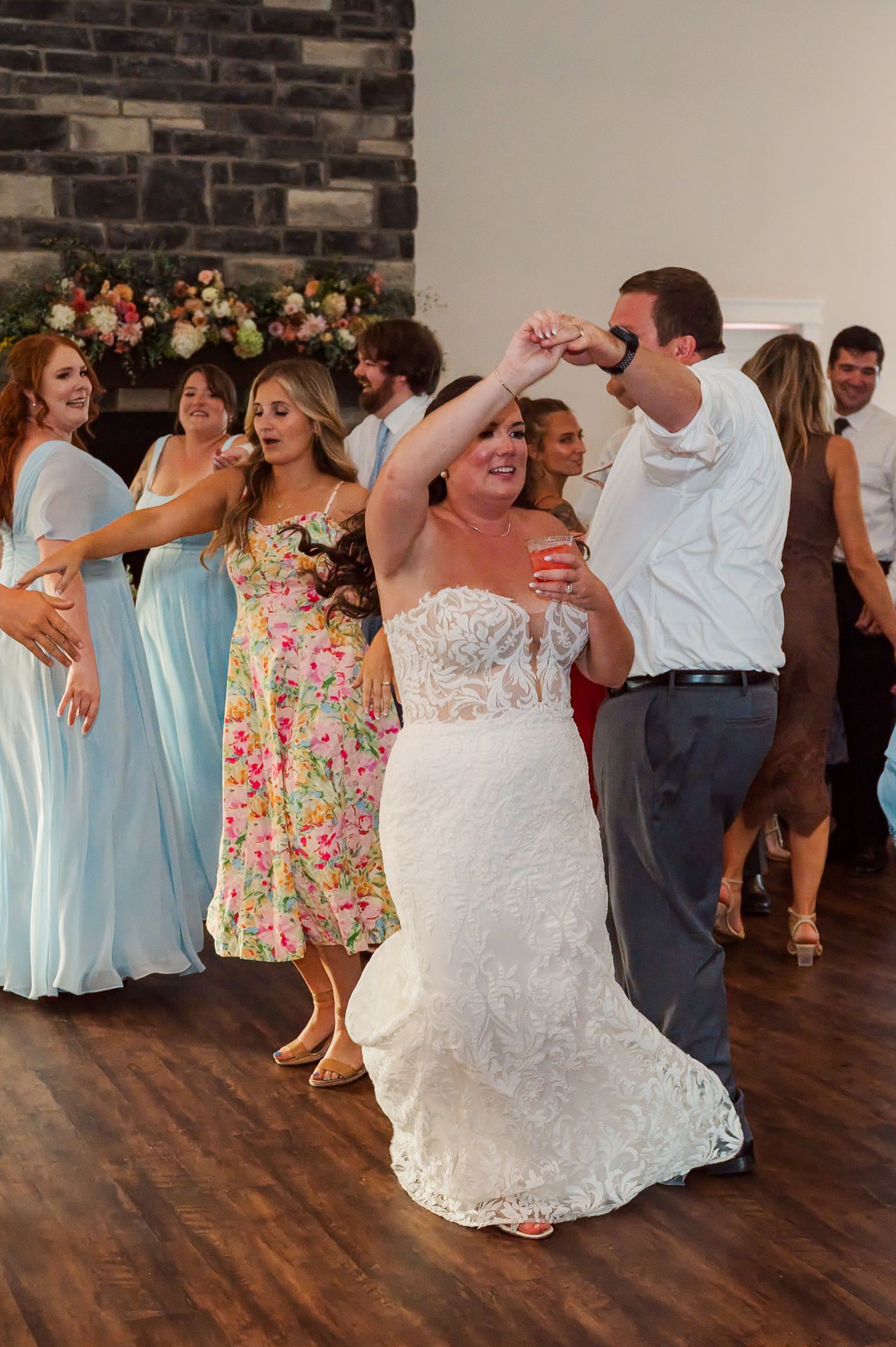 Bride in a white lace gown dances with a man at a wedding reception while bridesmaids in blue and floral dresses celebrate nearby.