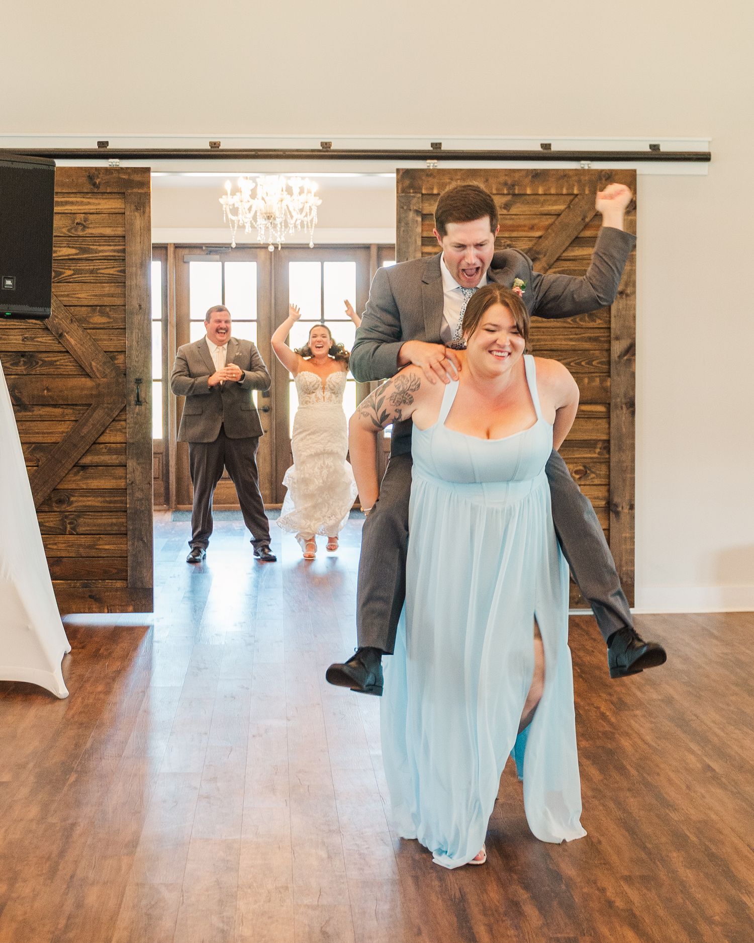 Groomsman carries bridesmaid through barn doors as bride and groom make a fun grand entrance at reception.
