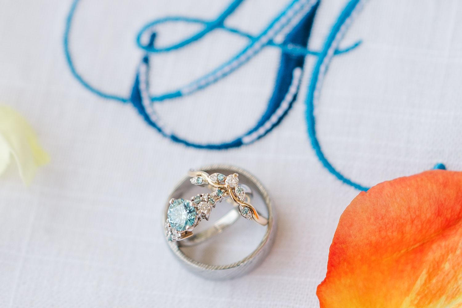 Elegant wedding rings with aquamarine and floral gold details on white linen with blue monogram embroidery.