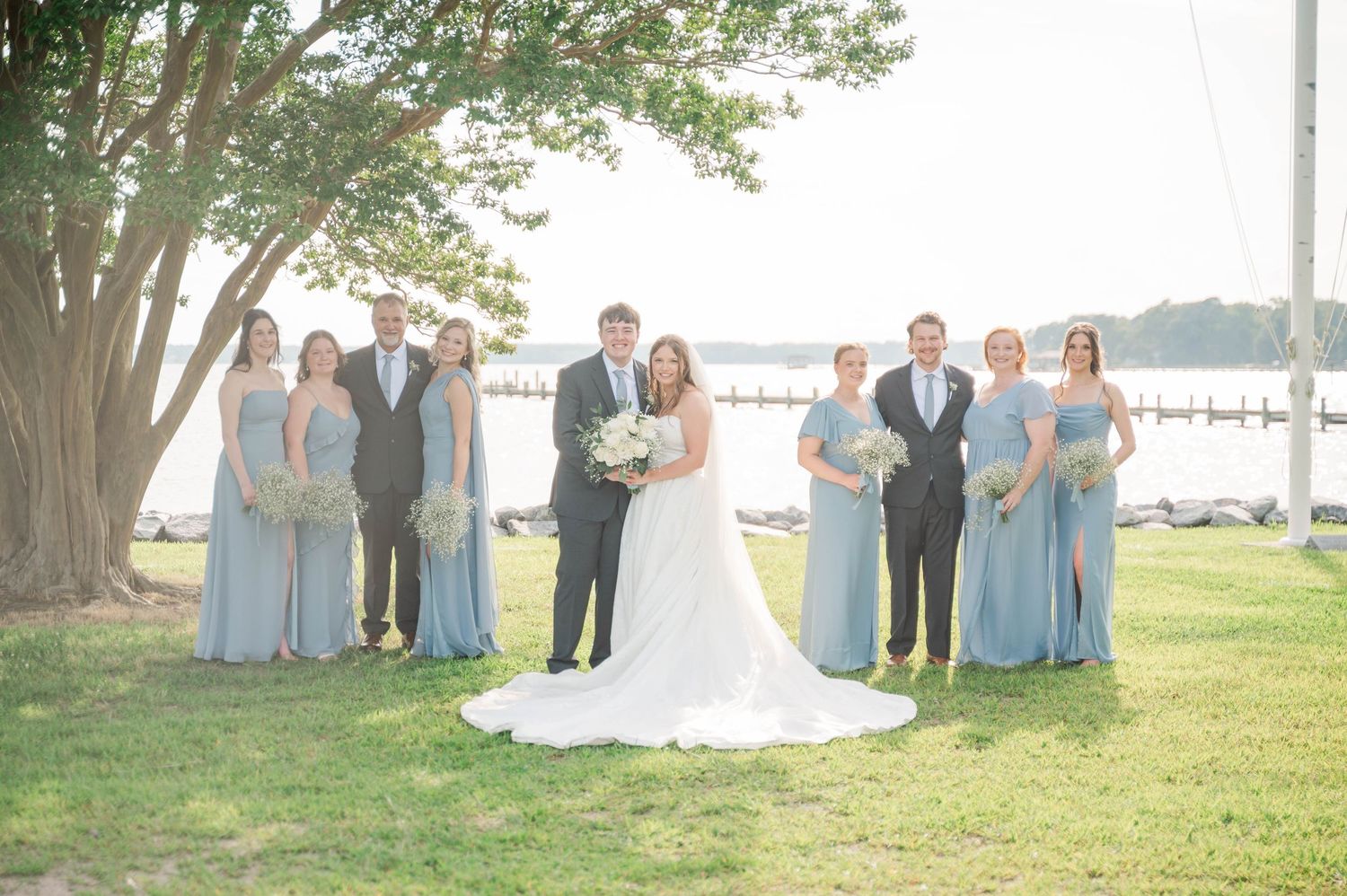 Wedding group photo with bridesmaids in blue dresses arranged under a shady tree near the water.