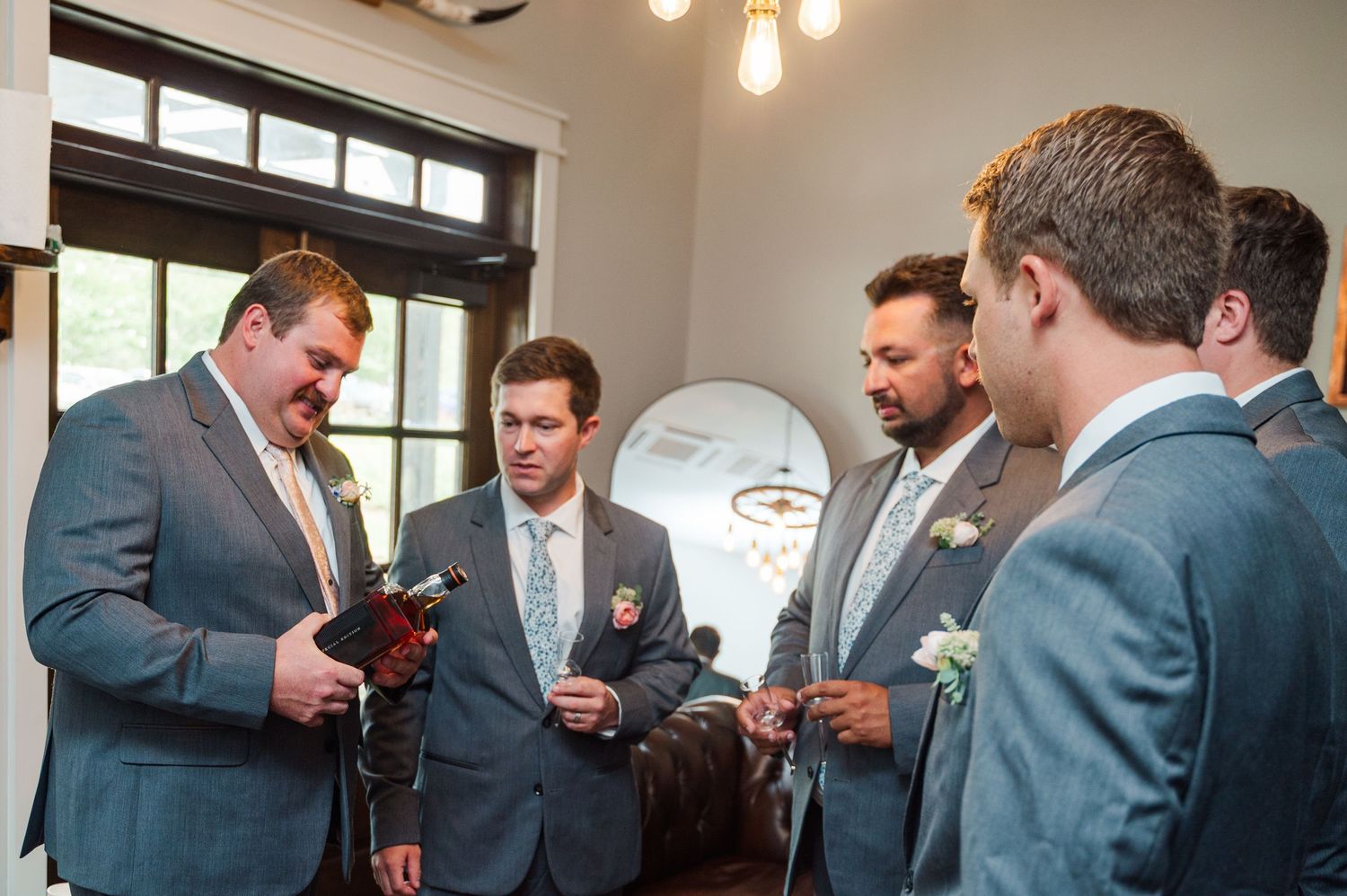 Groomsmen in gray suits with boutonnieres gather indoors before a wedding ceremony.