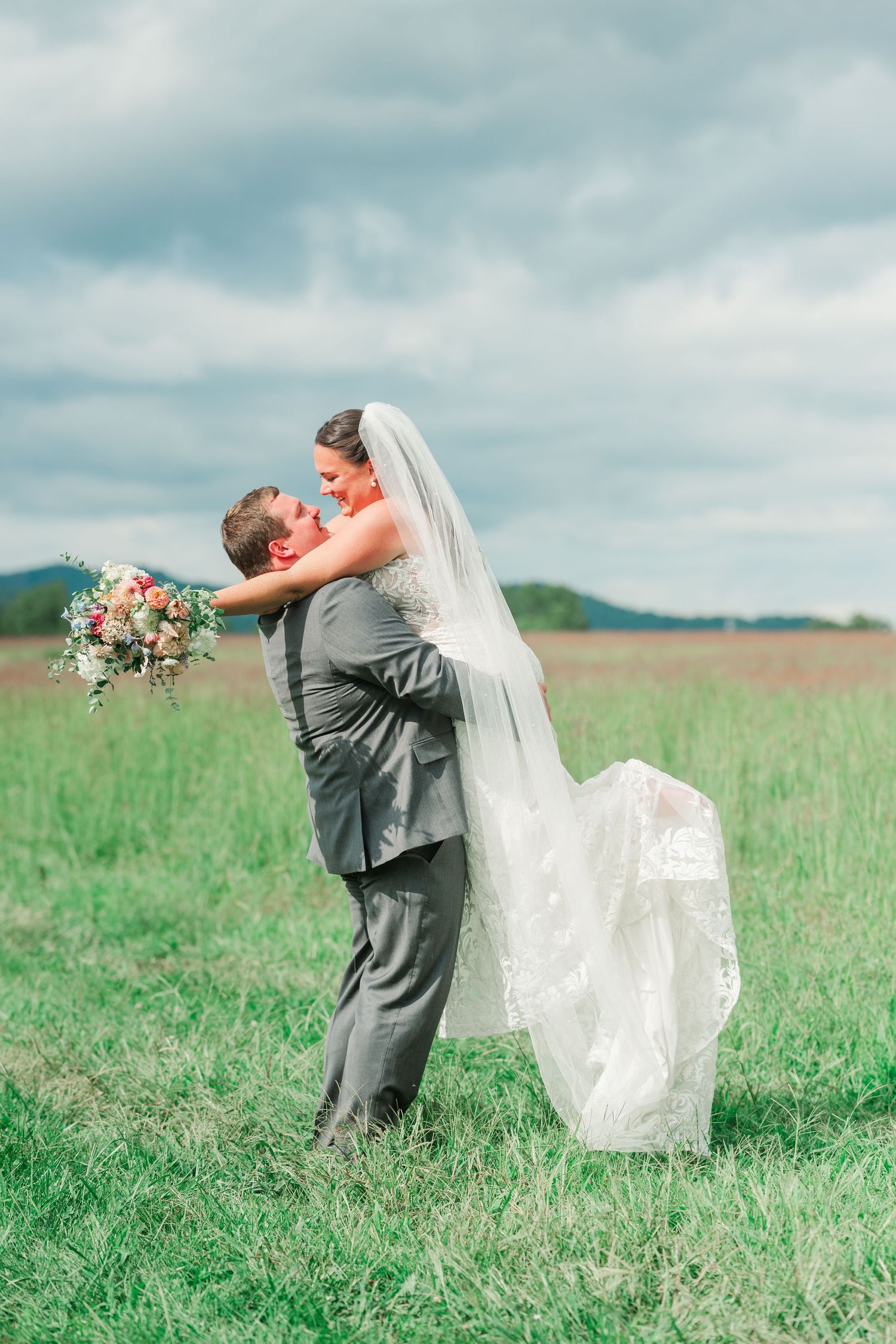 Groom lifts bride in white lace gown and veil for a kiss in a green field under cloudy skies.