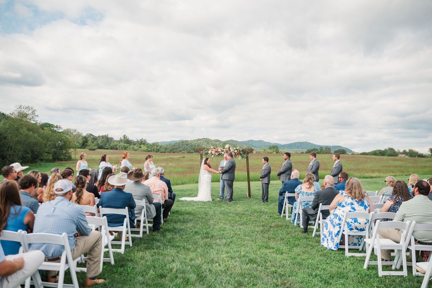 Outdoor wedding ceremony on green grass with guests seated in white chairs, floral arch, and mountain backdrop.