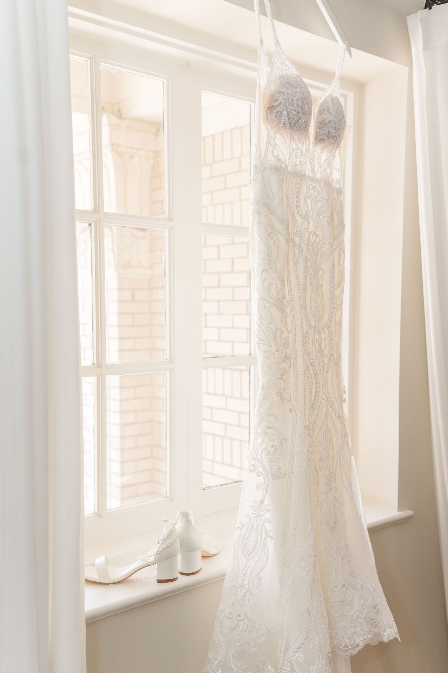 Wedding dresses hanging in bright window light with white curtains.