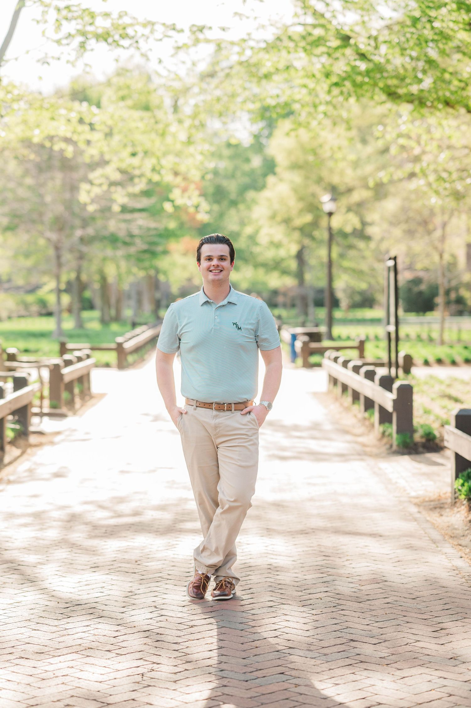 A person in a mint striped polo and khakis walks along a brick pathway surrounded by greenery.