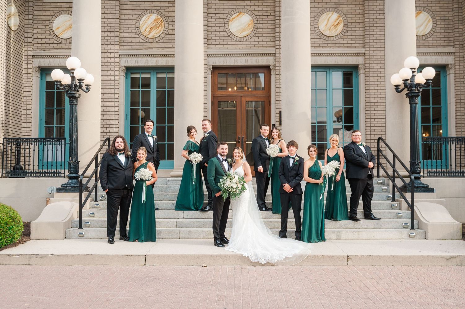 Wedding party in emerald green gowns and black tuxedos posed on steps of brick building with bride and groom centered.