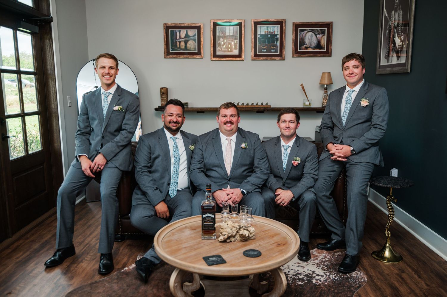 Five groomsmen in gray suits smiling together in an elegant room with framed artwork, gathered around a wooden coffee table.