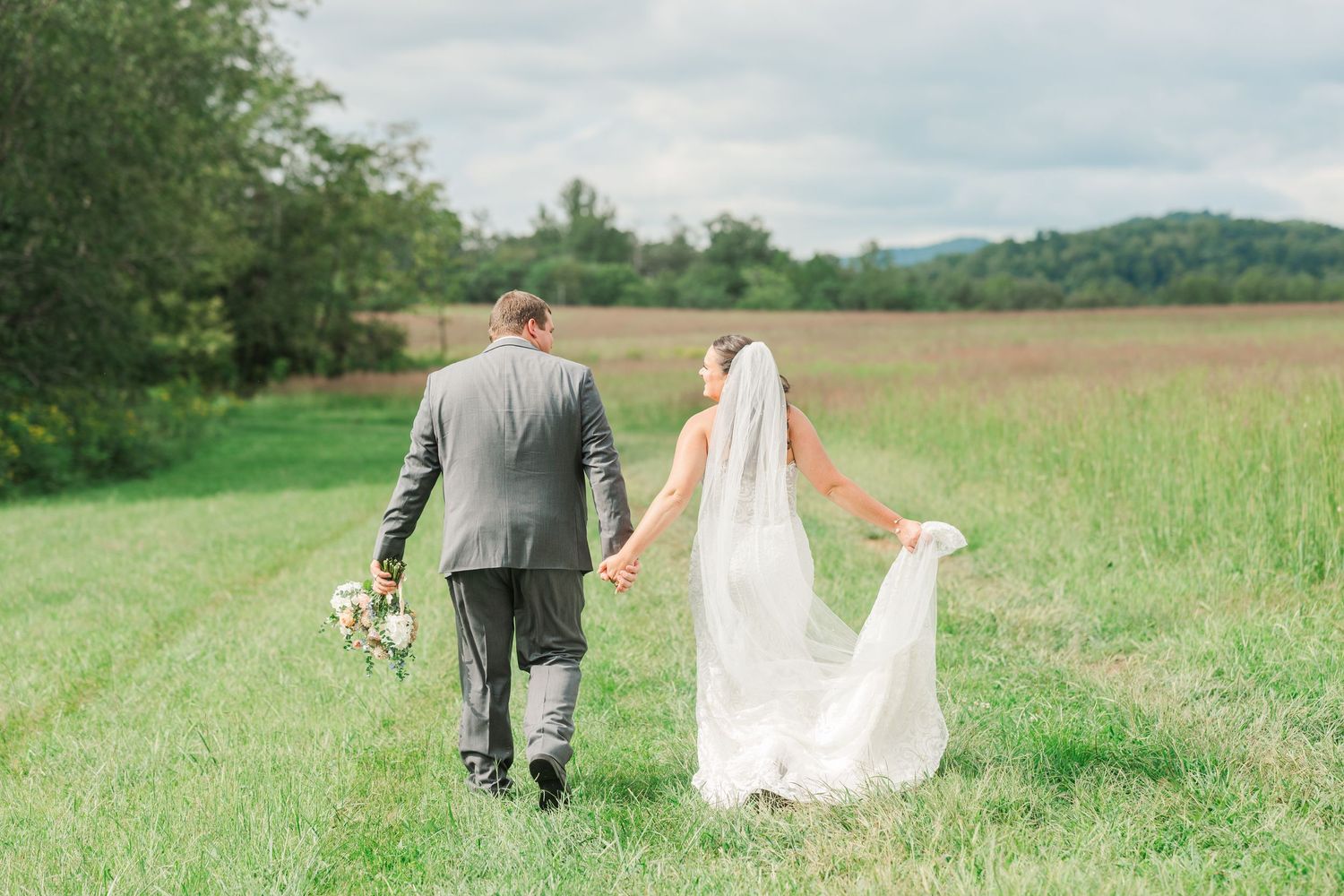 Bride and groom walk hand-in-hand through a lush green field with rolling hills and trees in the background.