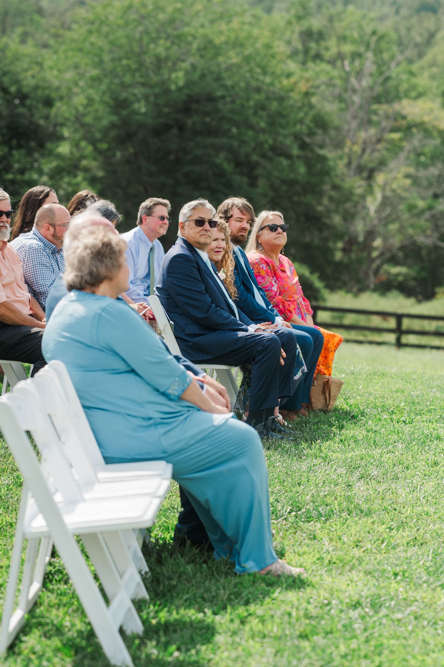 Wedding guests seated in white chairs on grass watching an outdoor ceremony on a sunny day.