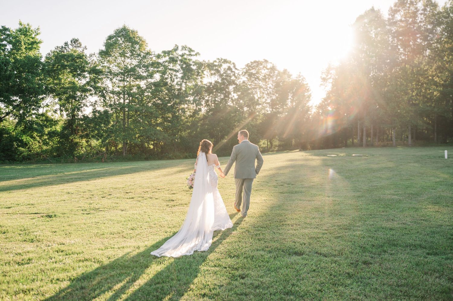 Bride and groom walking hand in hand across a sunlit green field with golden hour rays shining through trees.