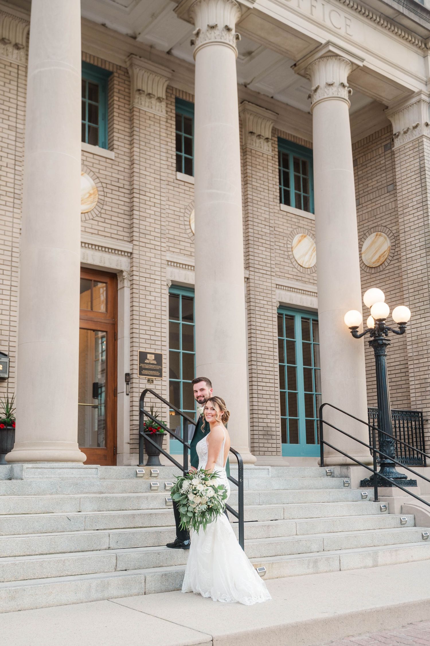 A wedding couple stands on grand white stone steps with classic columns in the background.