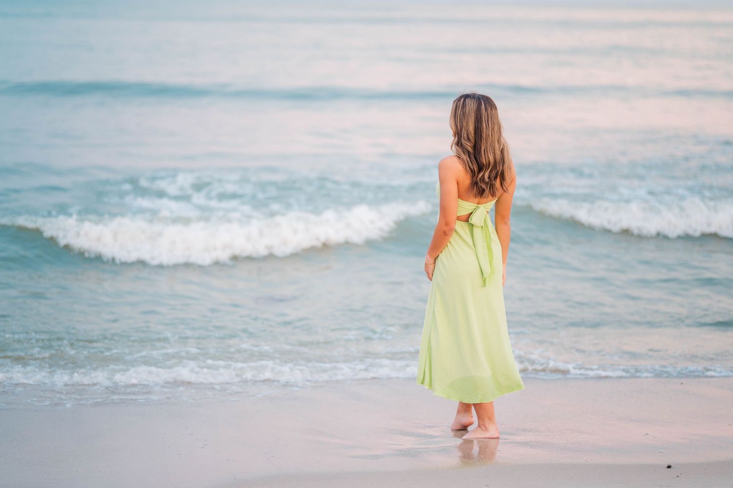 A woman in a light green maxi dress stands barefoot on a sandy beach, gazing at gentle ocean waves.