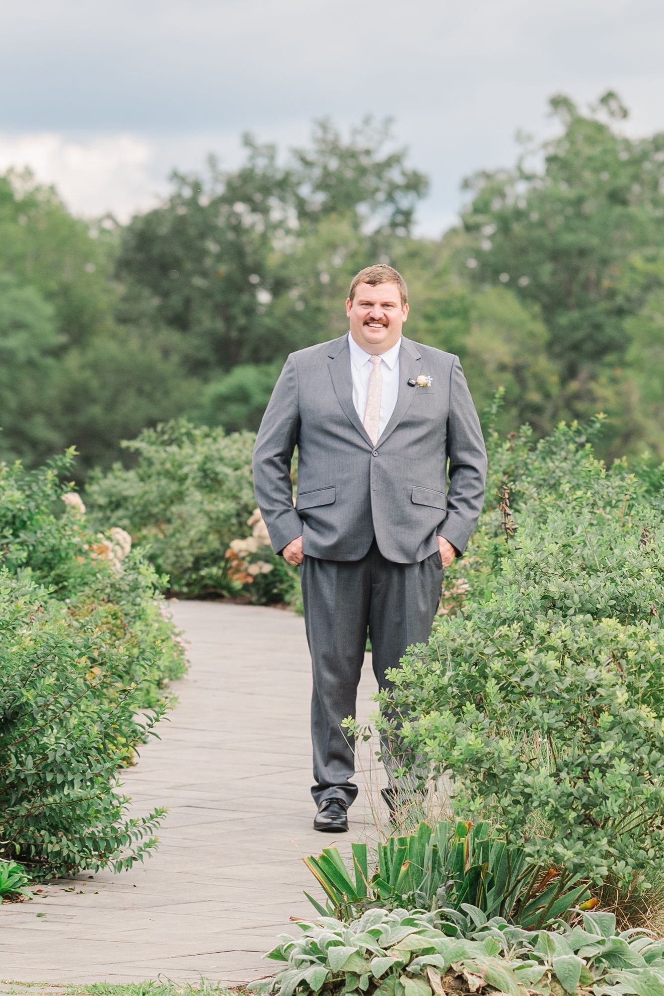 Groom in gray suit walks garden path lined with lush greenery, smiling confidently on his wedding day.
