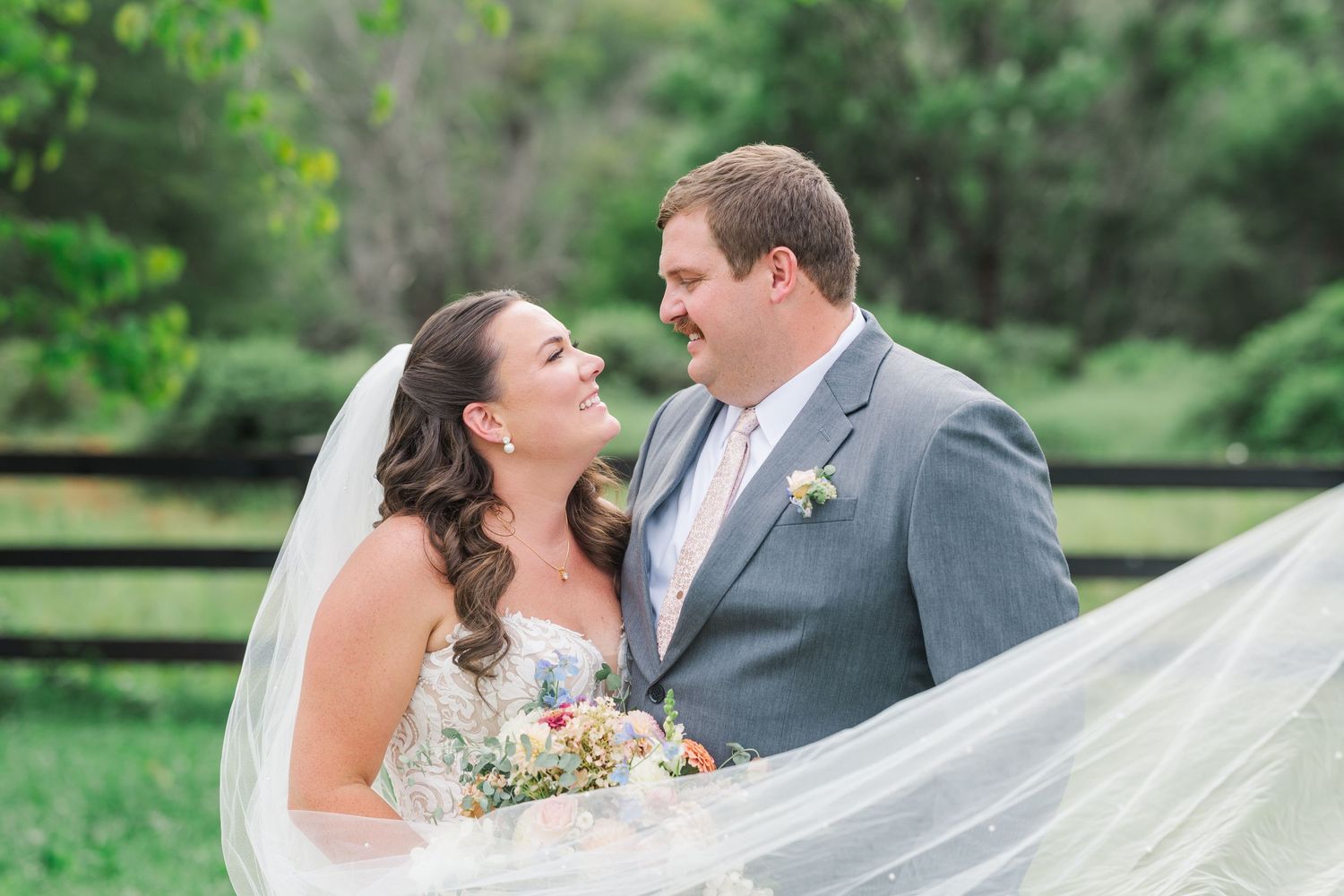 Bride and groom smiling at each other outdoors with wedding veil flowing in the breeze near a green fence.