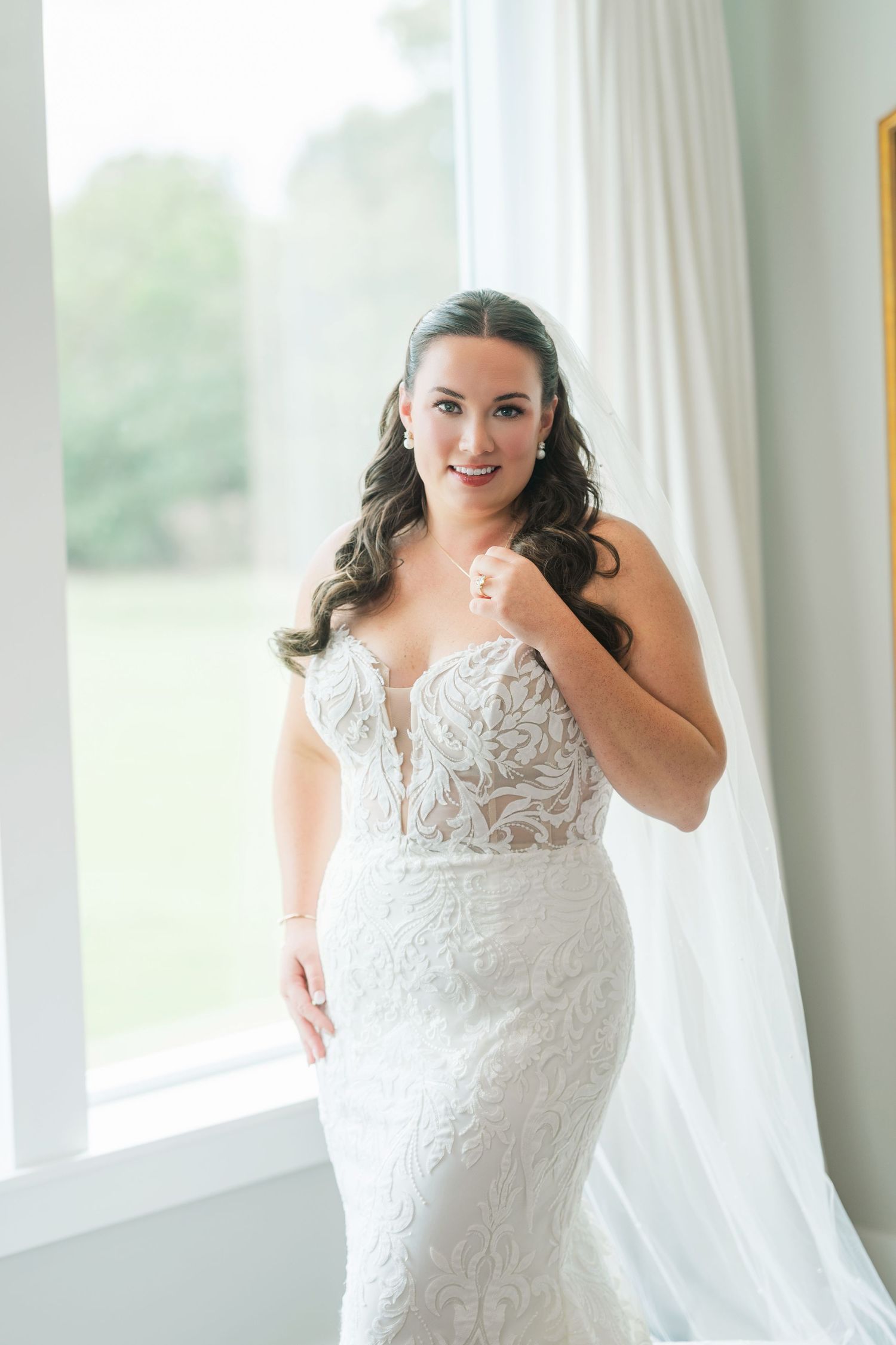 Bride in lace mermaid wedding gown poses by window with flowing veil, smiling elegantly on her wedding day.