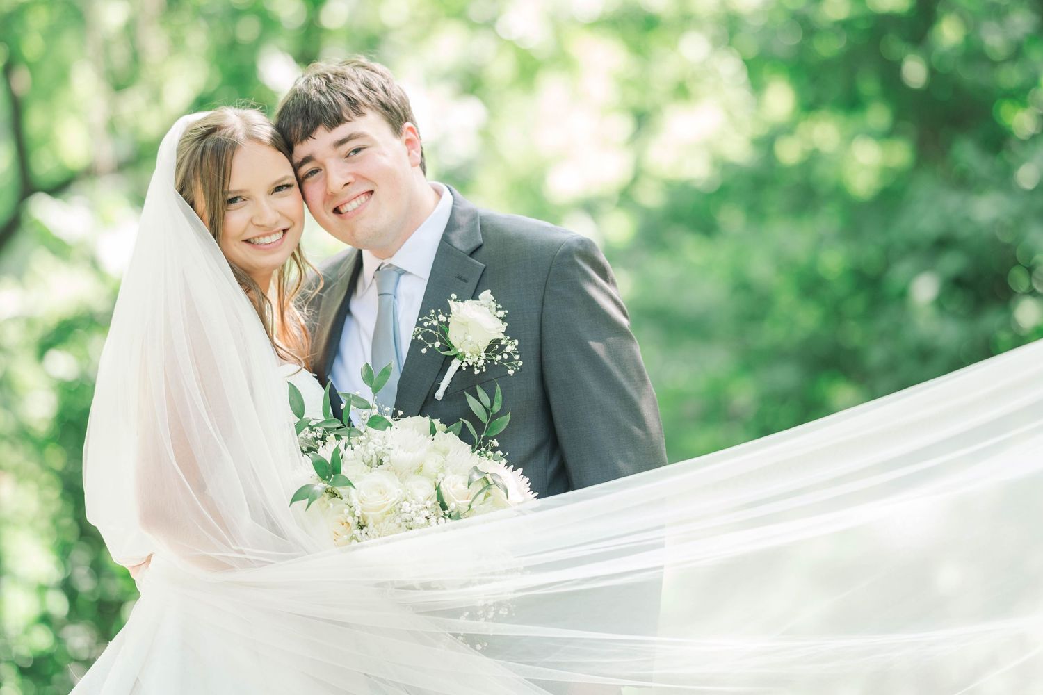 Romantic wedding couple embraces in a lush green garden setting with flowing veil and white floral bouquet.