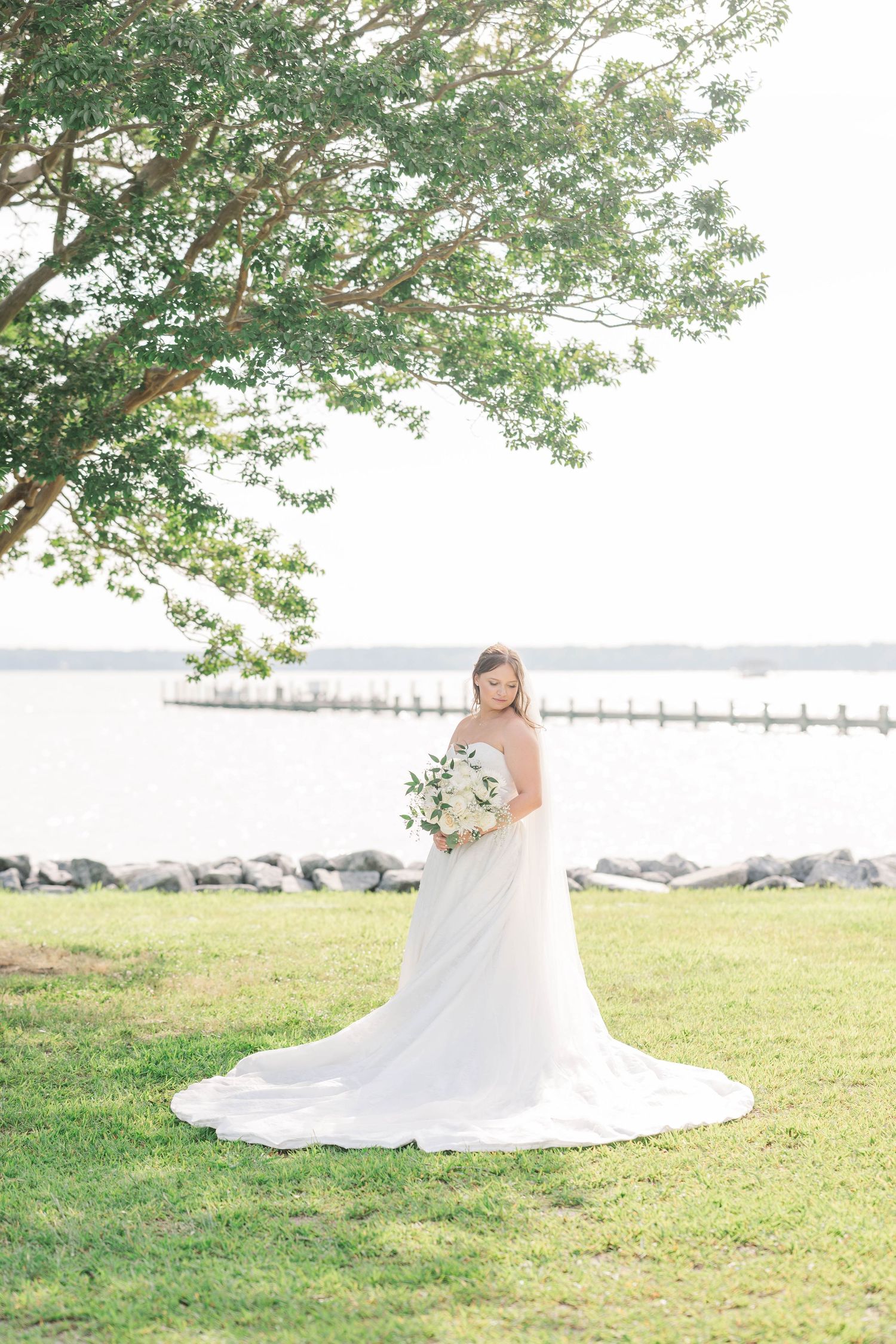 Elegant bride in flowing white gown poses on a hillside with water and dock in the background.