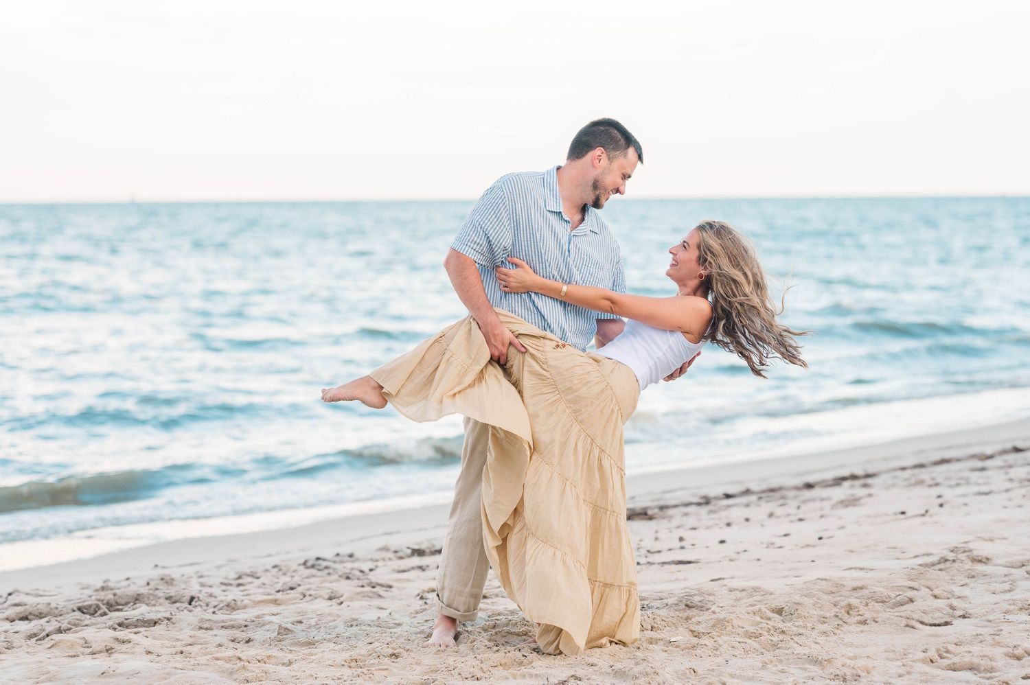 A couple in beige attire walks along a sandy beach with ocean waves in the background.