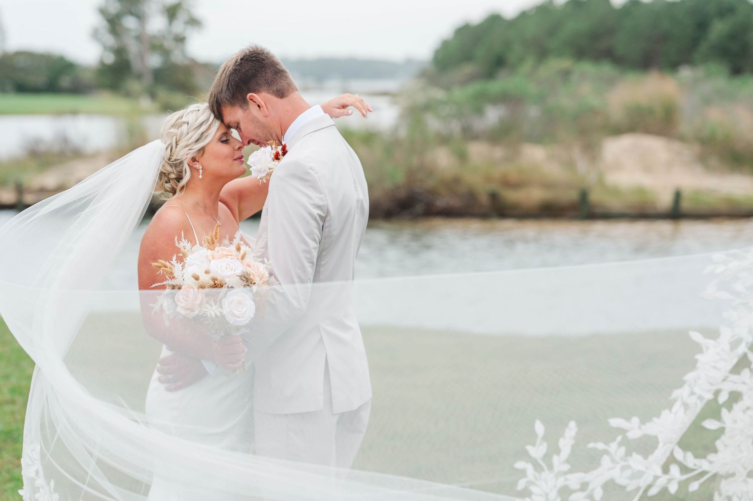 Bride and groom embrace near waterfront, her lace veil billowing in wind, as she holds white bouquet in outdoor wedding portrait.