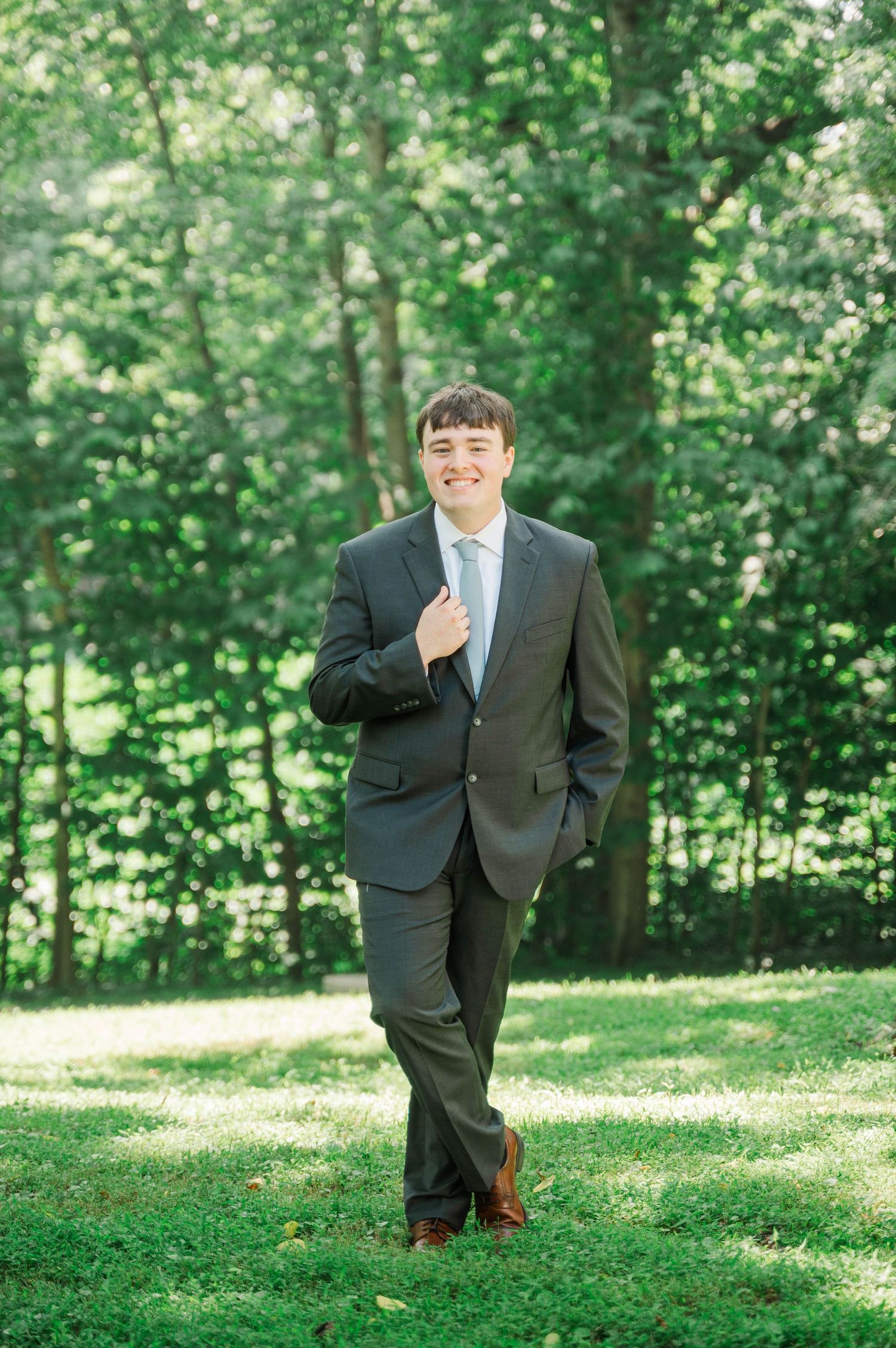Person in dark suit poses playfully on green grass in wooded setting.
