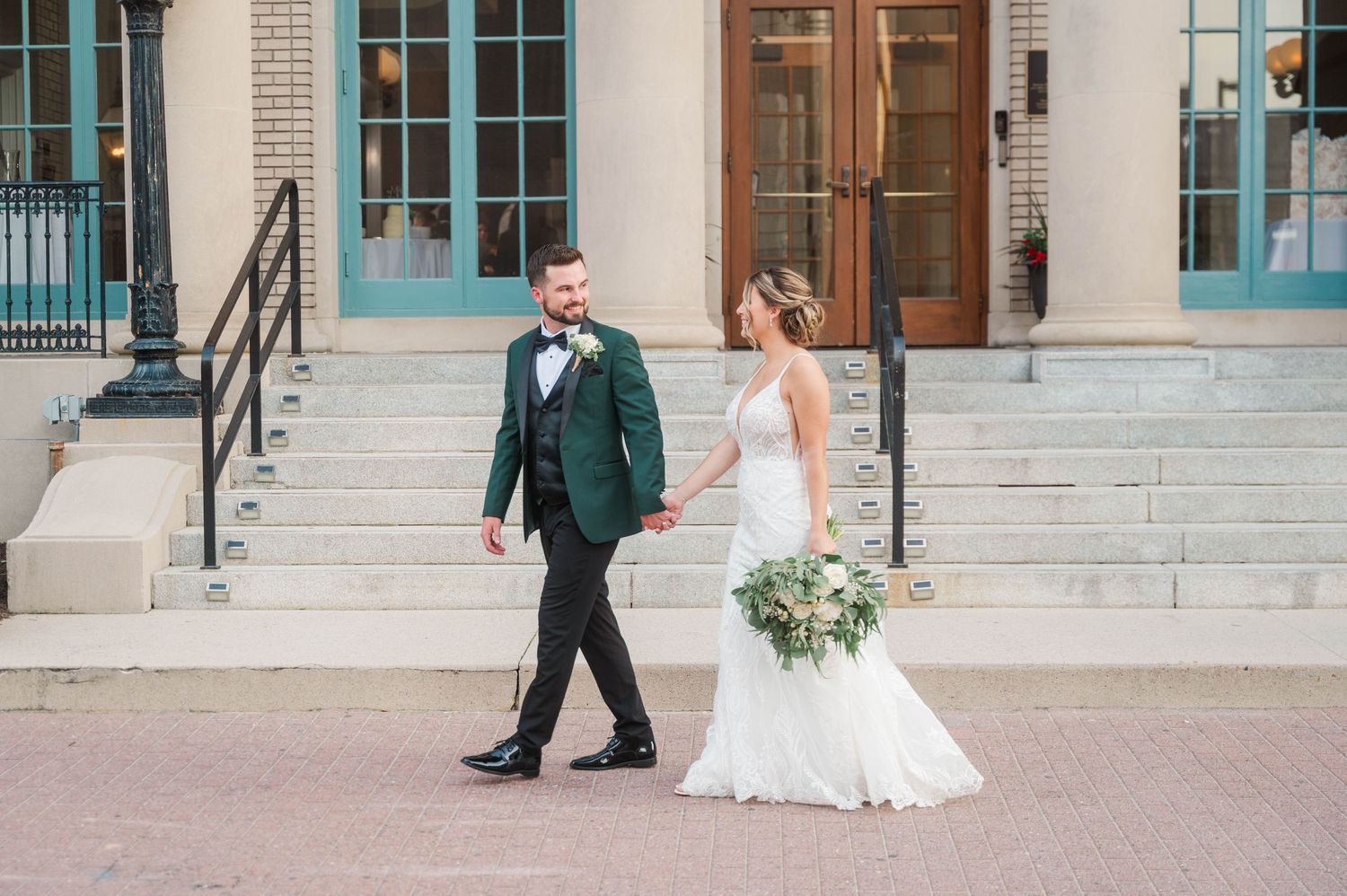 A couple walks along a classic brick building with white columns during their wedding day.