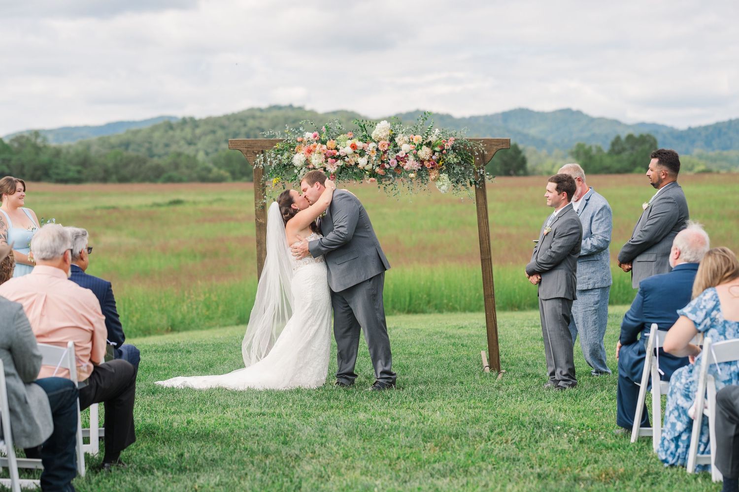 Bride and groom share their first kiss under a floral wooden arch with rolling green mountains in the background.