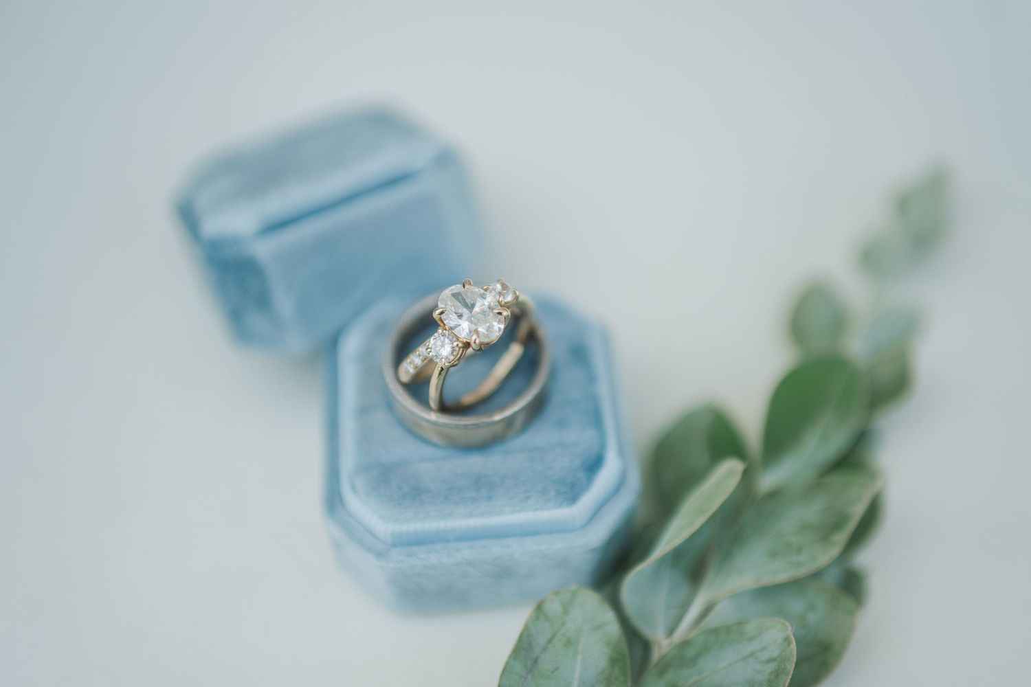 Diamond engagement ring and gold wedding band displayed in a blue velvet ring box with eucalyptus greenery.