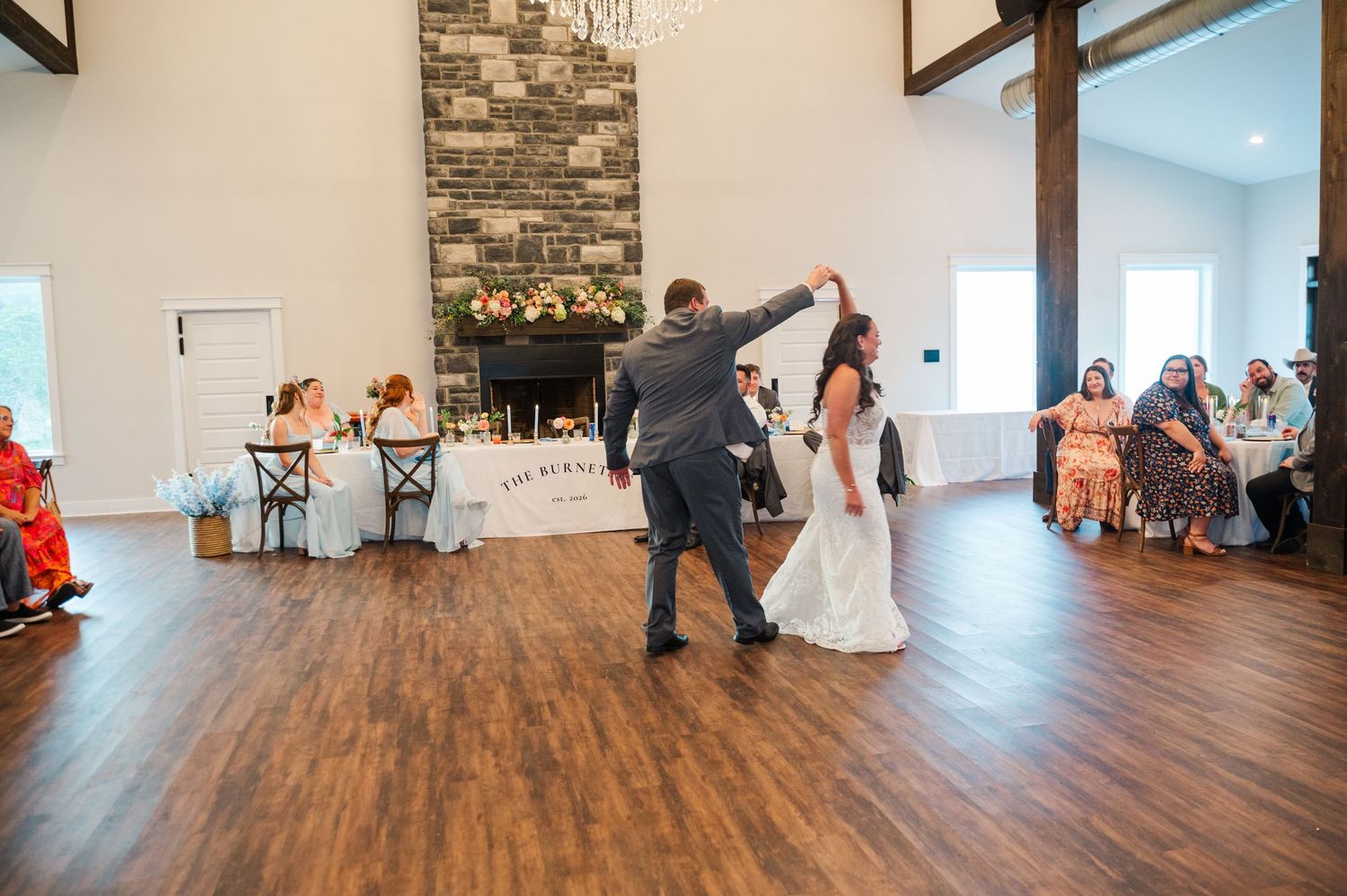 Bride in a white gown and groom in a gray suit share a first dance on a wood floor at a rustic wedding reception with guests watching.