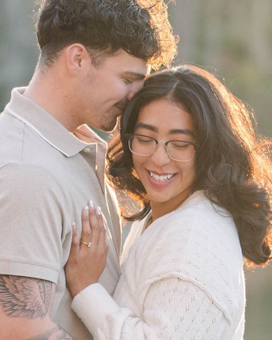 A smiling couple shares a tender moment outdoors, woman wearing glasses and ring, man with tattoo, bathed in golden light.