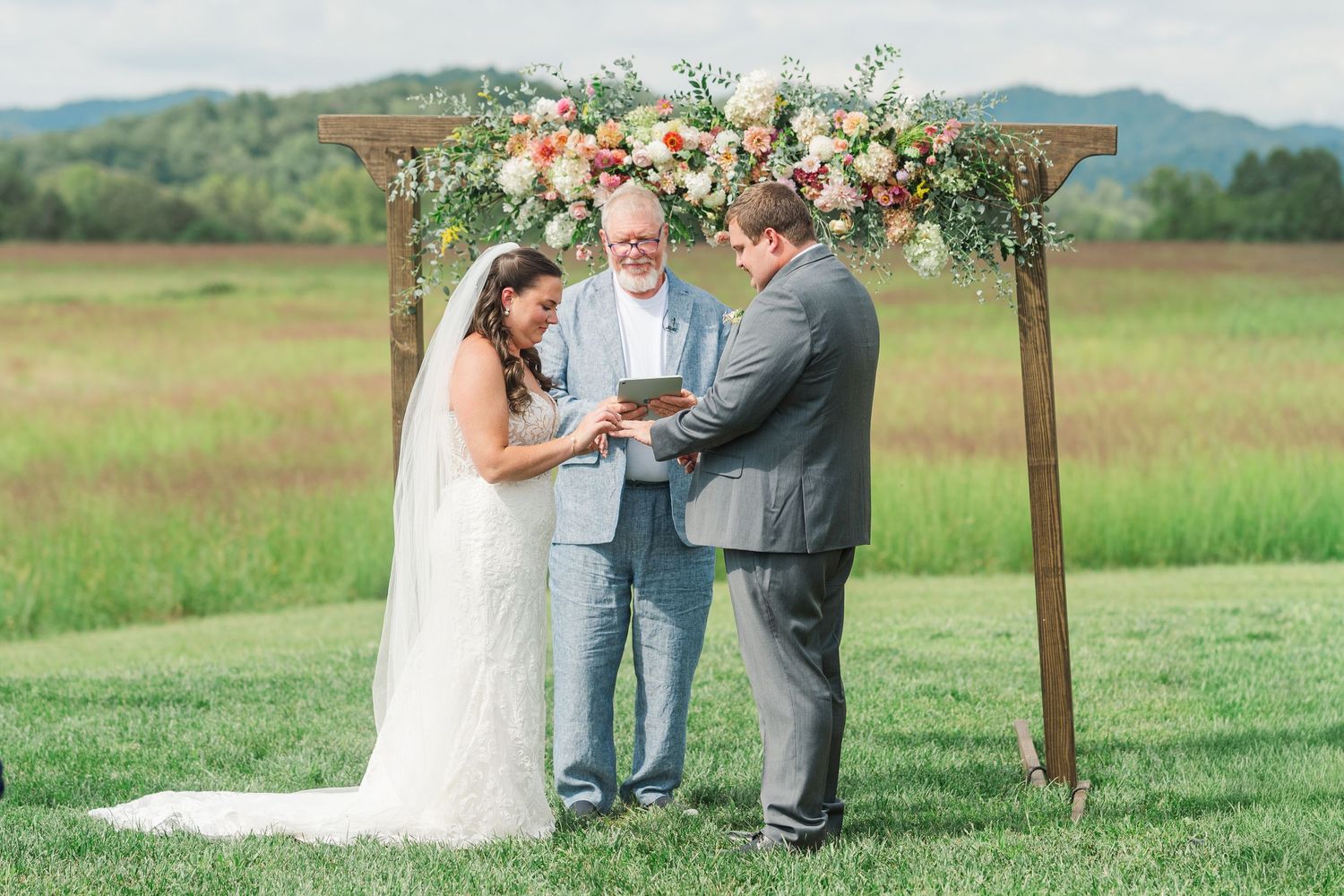 Outdoor wedding ceremony with bride and groom exchanging rings under a wooden arch adorned with peach and white floral arrangements.