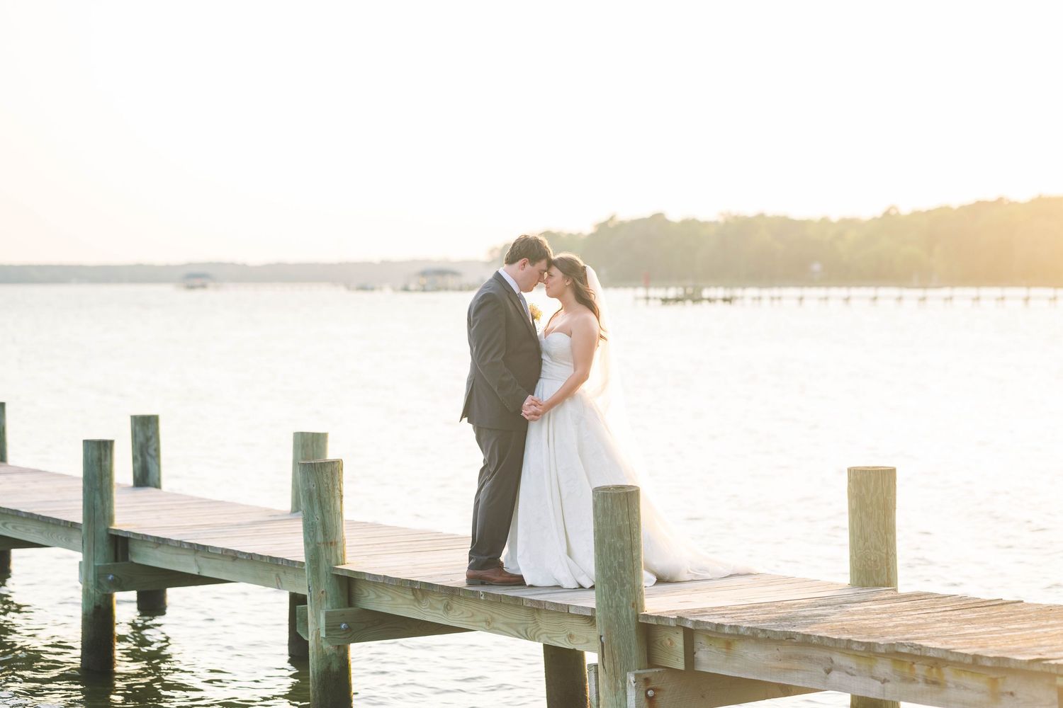 Newlyweds embrace on a waterfront pier during golden hour with boats in the distance.