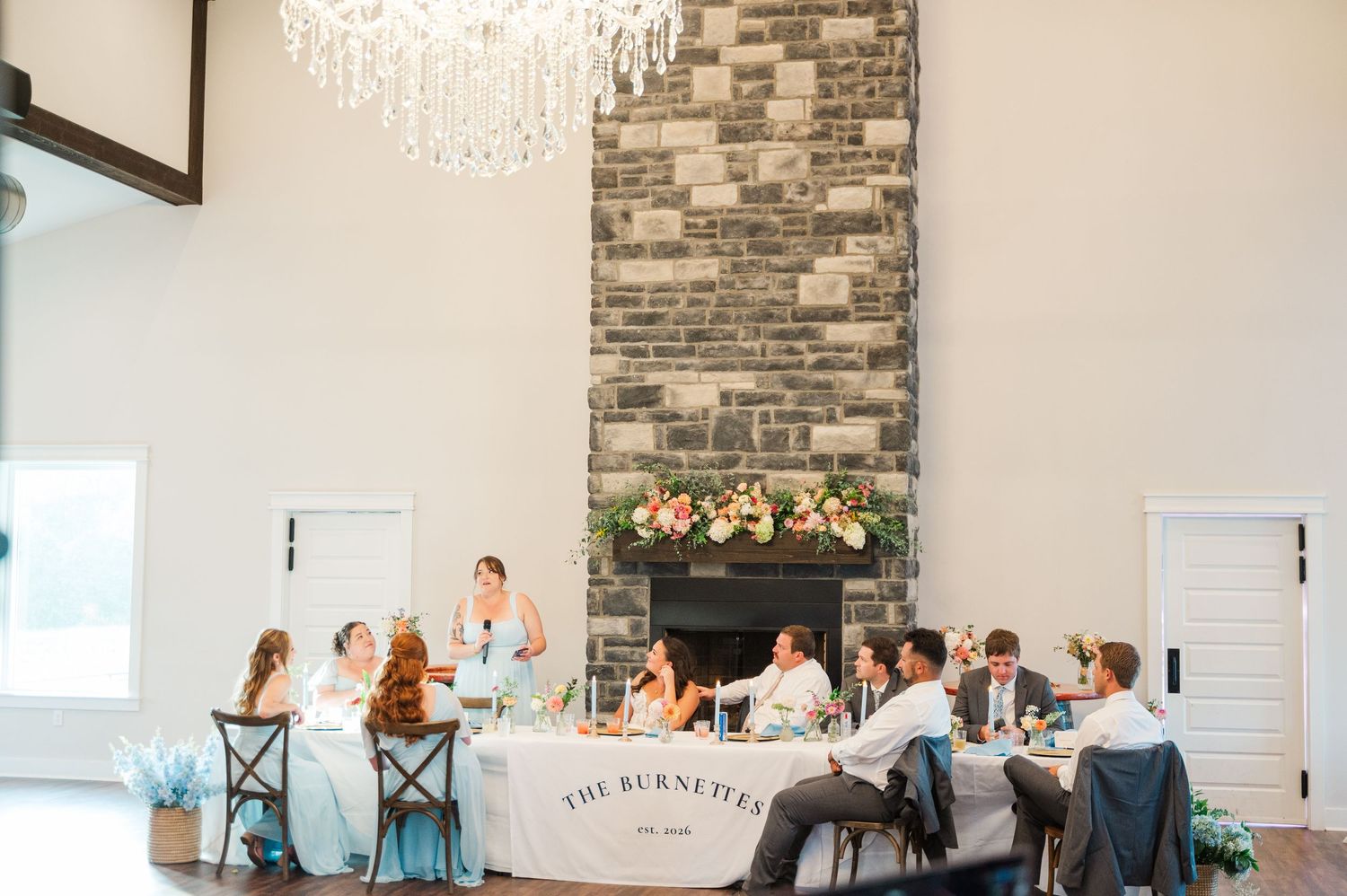 Wedding party seated at sweetheart table near stone fireplace under elegant crystal chandelier.