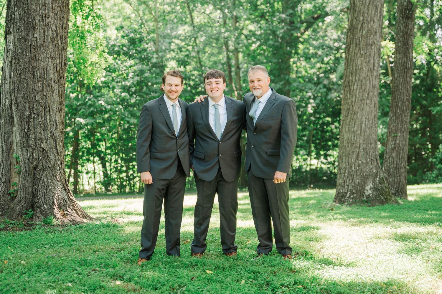 Wedding party members in dark suits posed casually in outdoor woodland setting.
