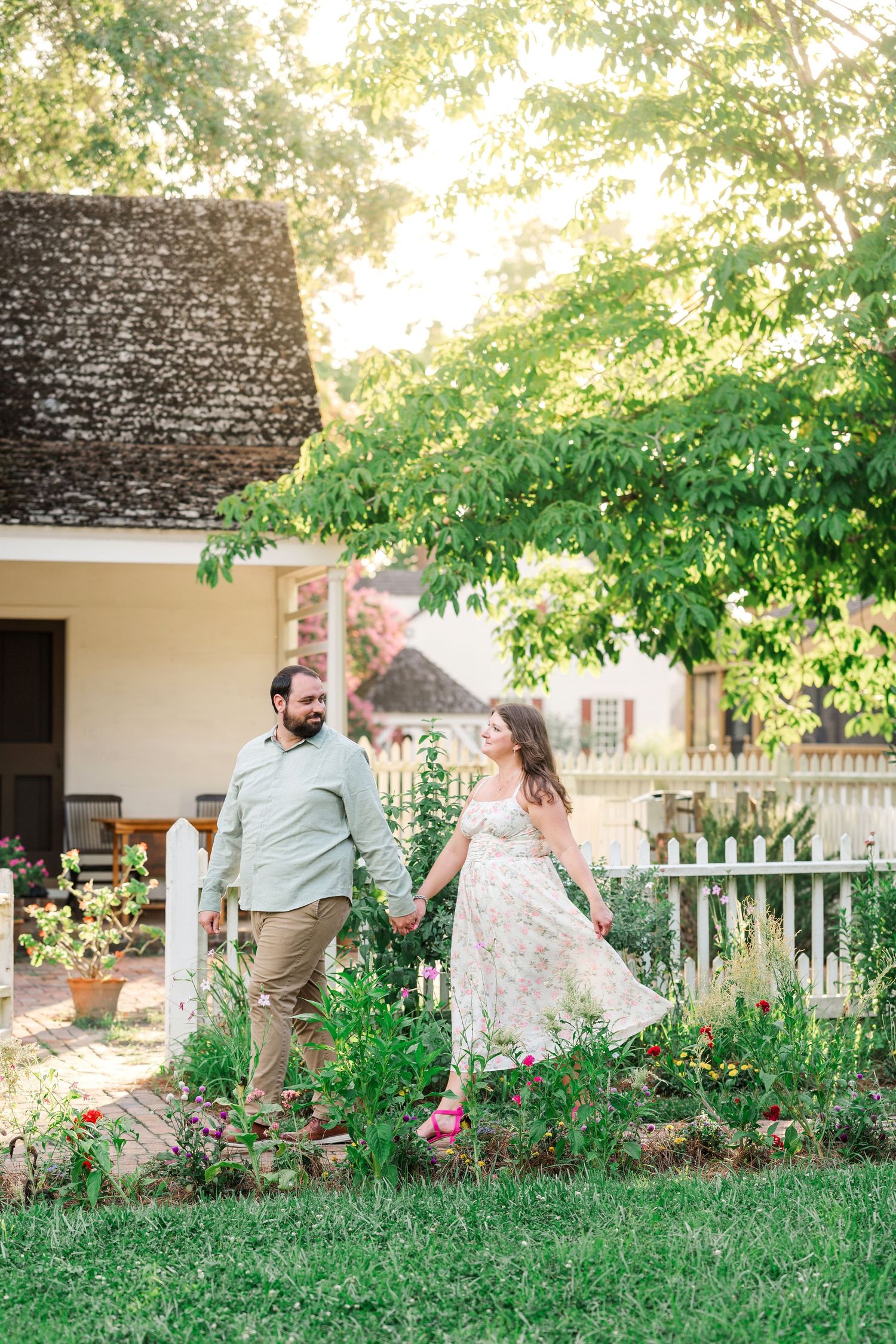 Couple holding hands in a lush garden beside a white picket fence and historic cottage at golden hour. Engagement. Williamsburg