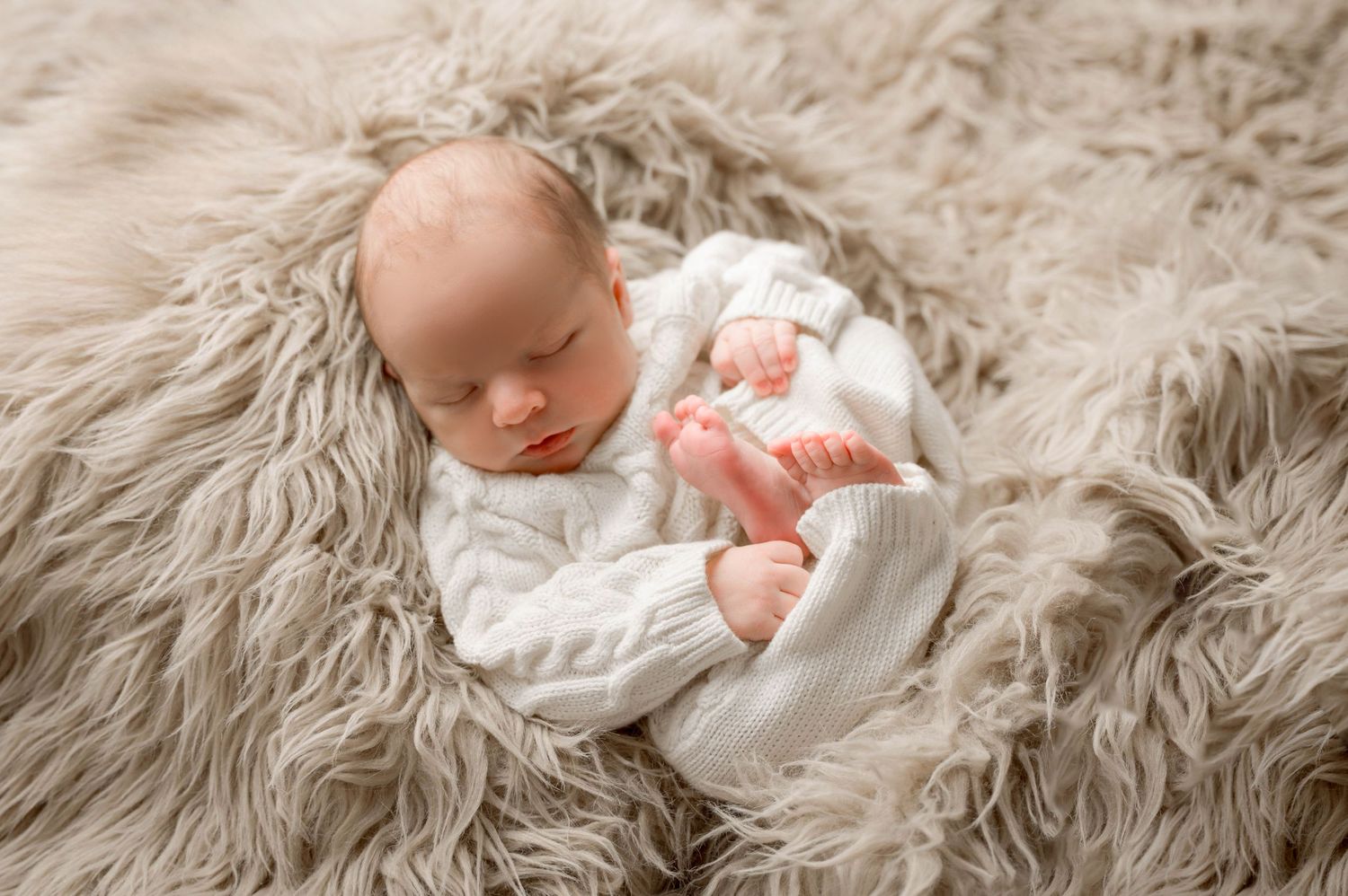 Peaceful newborn baby sleeping on fluffy beige fur blanket wearing white knit sweater in professional photography.