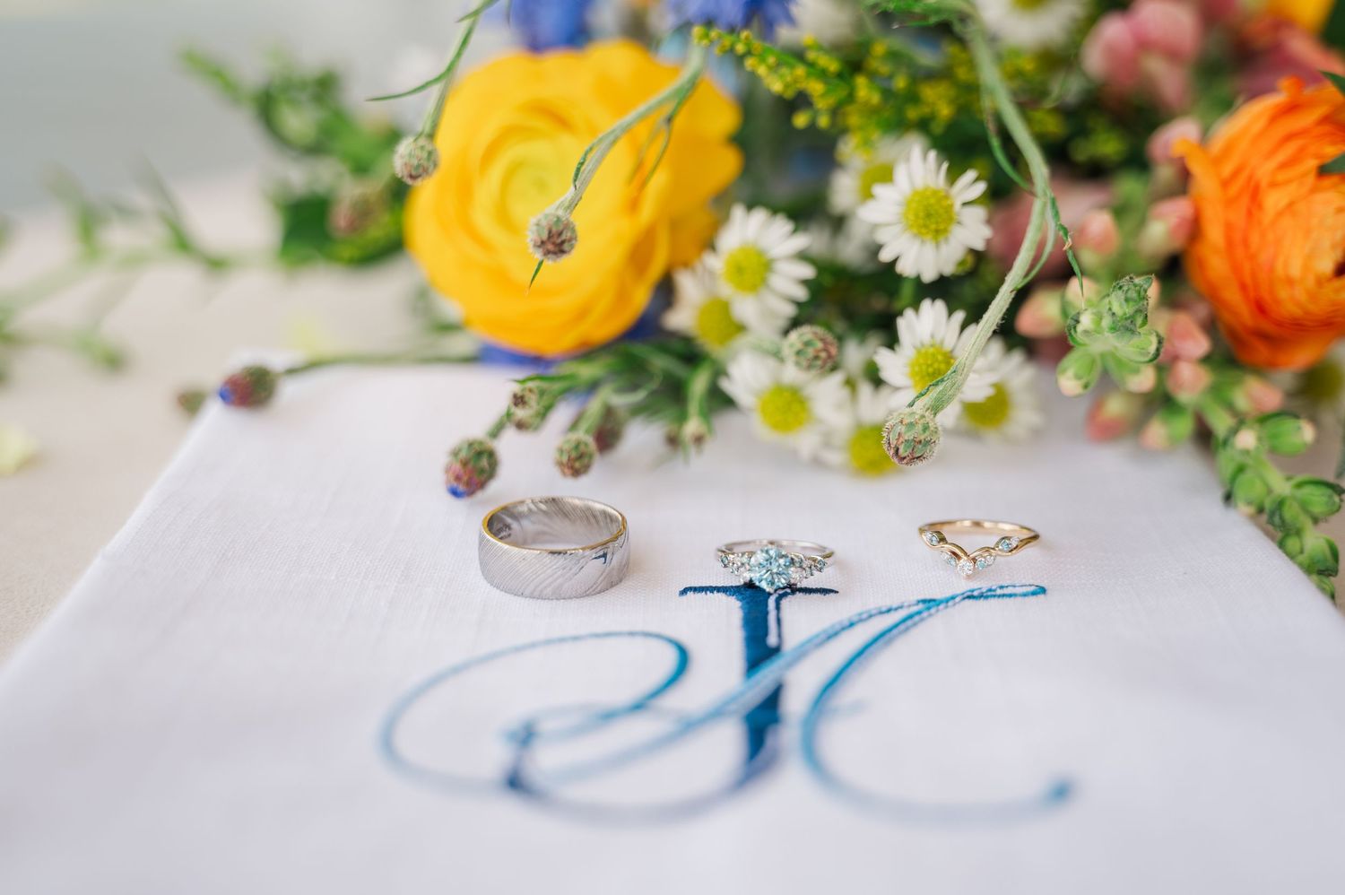 Wedding rings and diamond engagement ring on monogrammed white cloth with colorful floral bouquet backdrop.