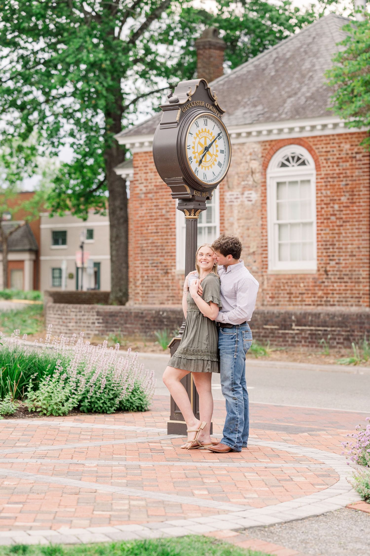 A couple embraces on a brick sidewalk near vintage street clocks and colonial architecture.