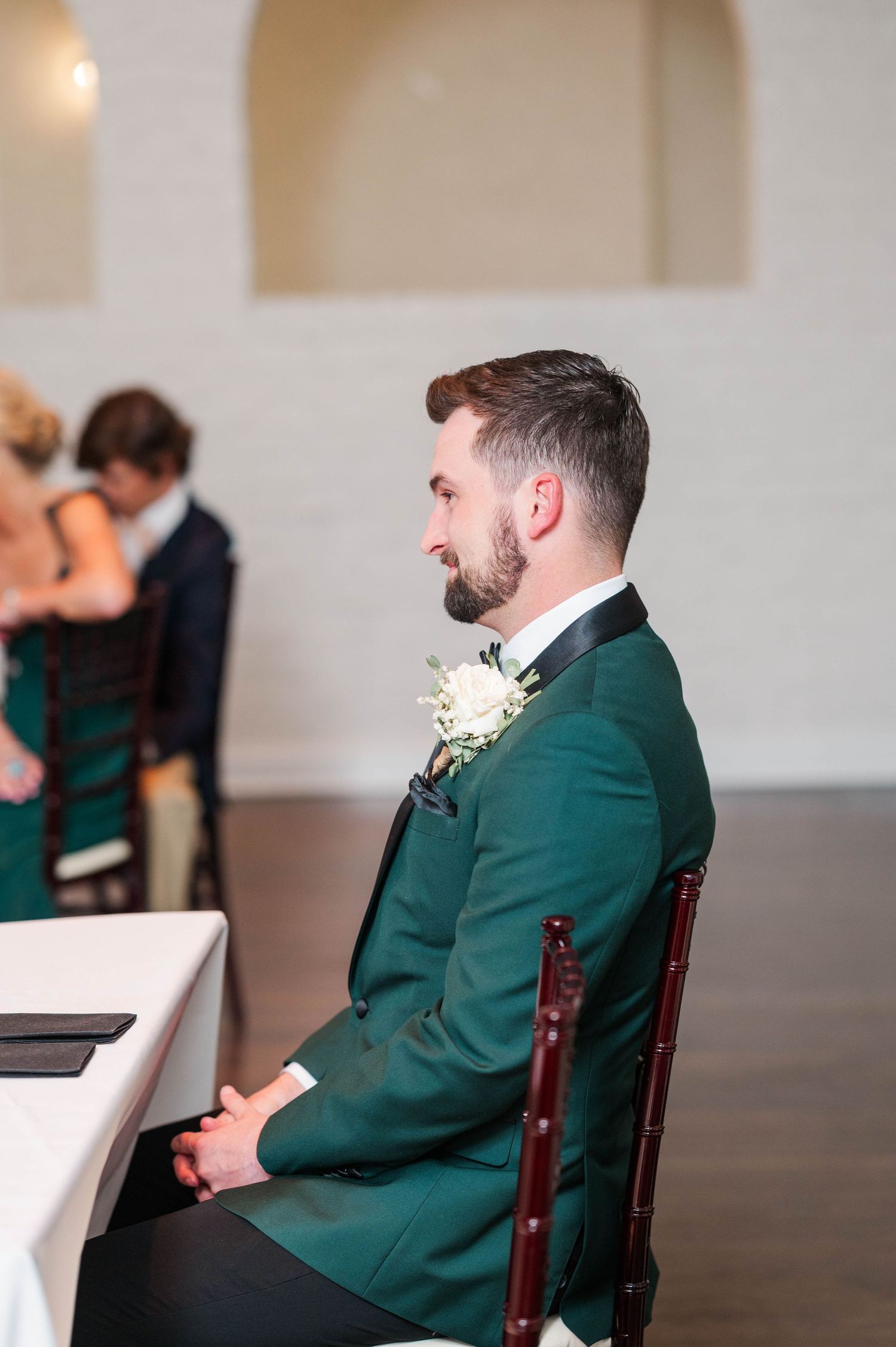 Groom in emerald green suit jacket seated during wedding reception.