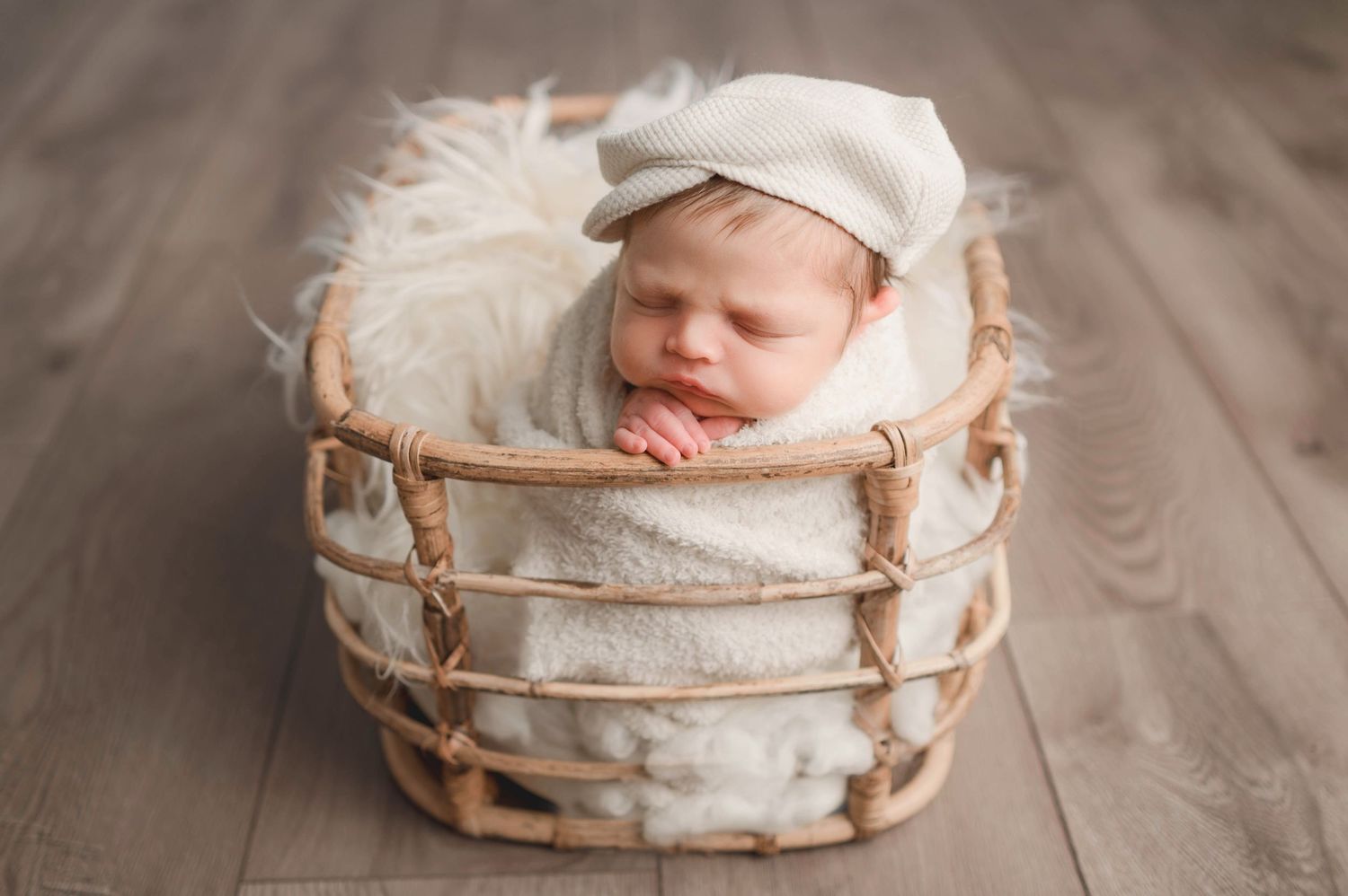 Sleeping newborn in white wrap nestled in rustic wooden basket on wooden floor.