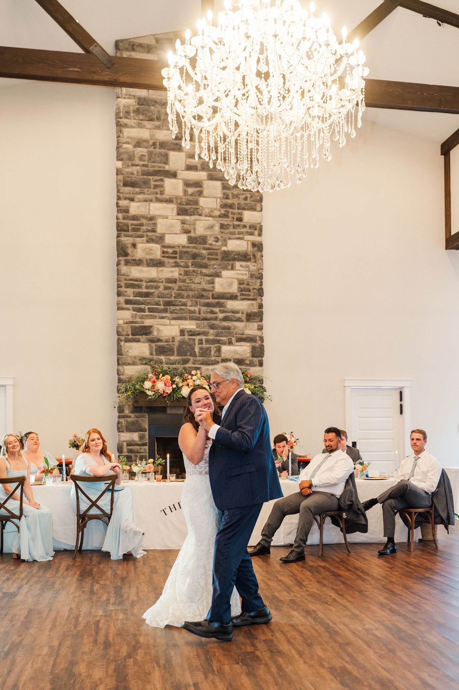 Bride shares an emotional father-daughter dance under a crystal chandelier in an elegant rustic ballroom.