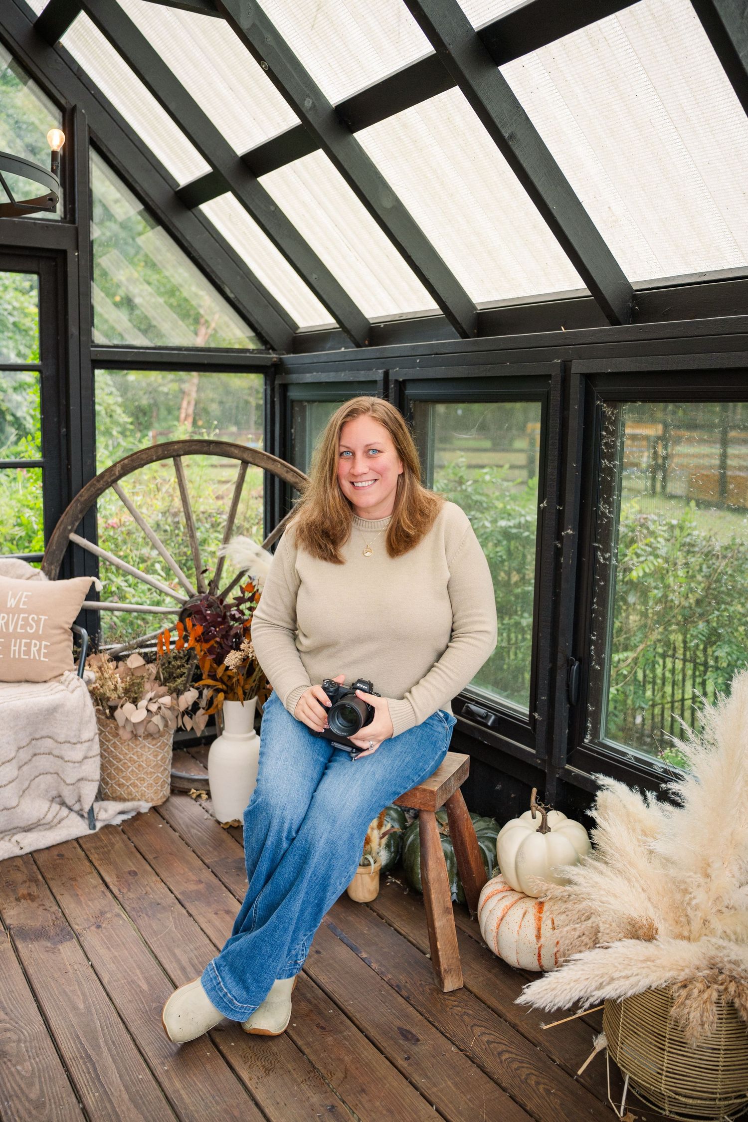 Photographer holding a camera sits on a wooden stool in a glass greenhouse with autumn pumpkins, pampas grass and a wagon wheel.